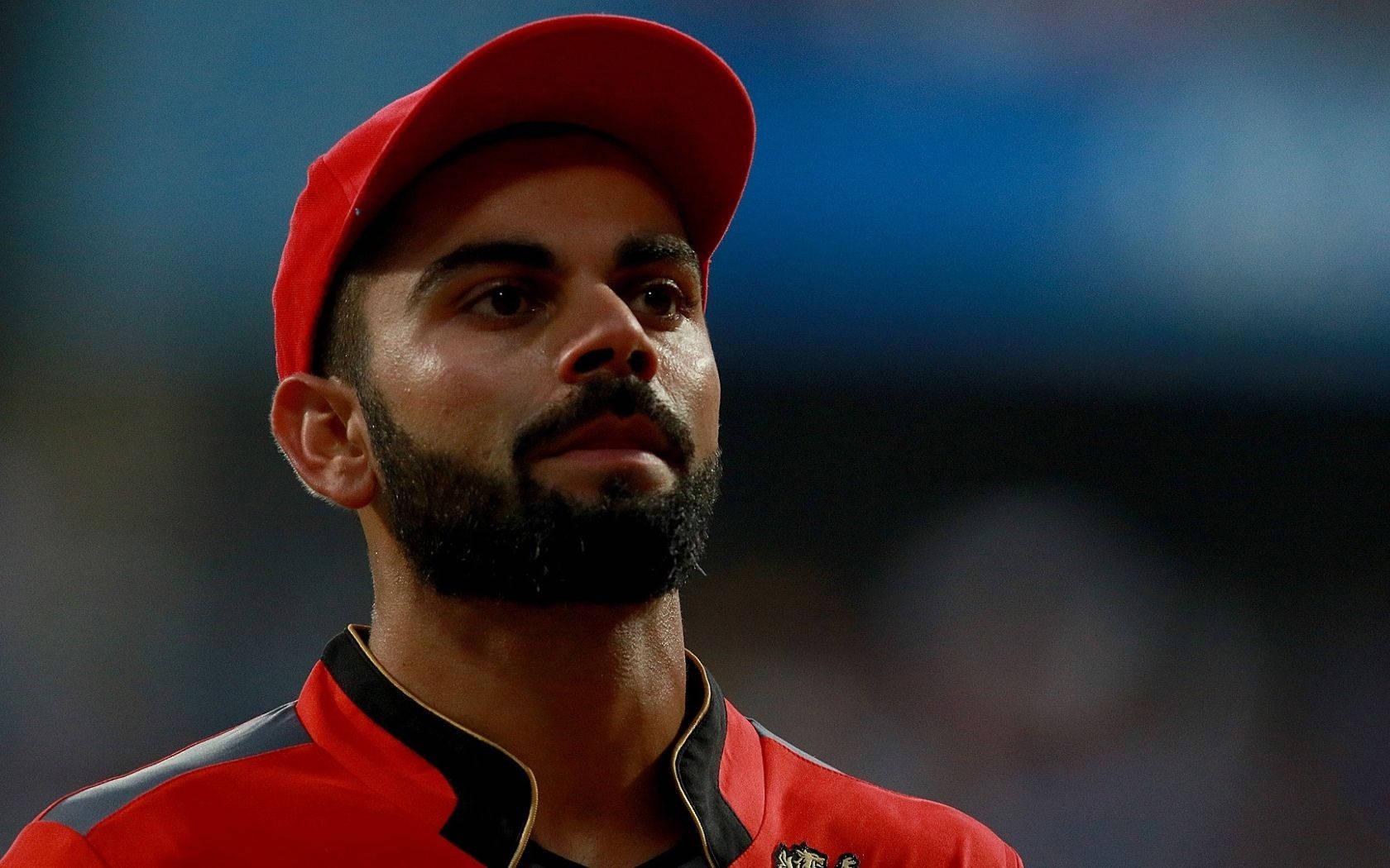 virat kohli rcb 桌布 壁紙 — 留鬍鬚的板球球員近距離肖像，戴著紅色帽子和球衣，在模糊的球場背景下專注向前看。