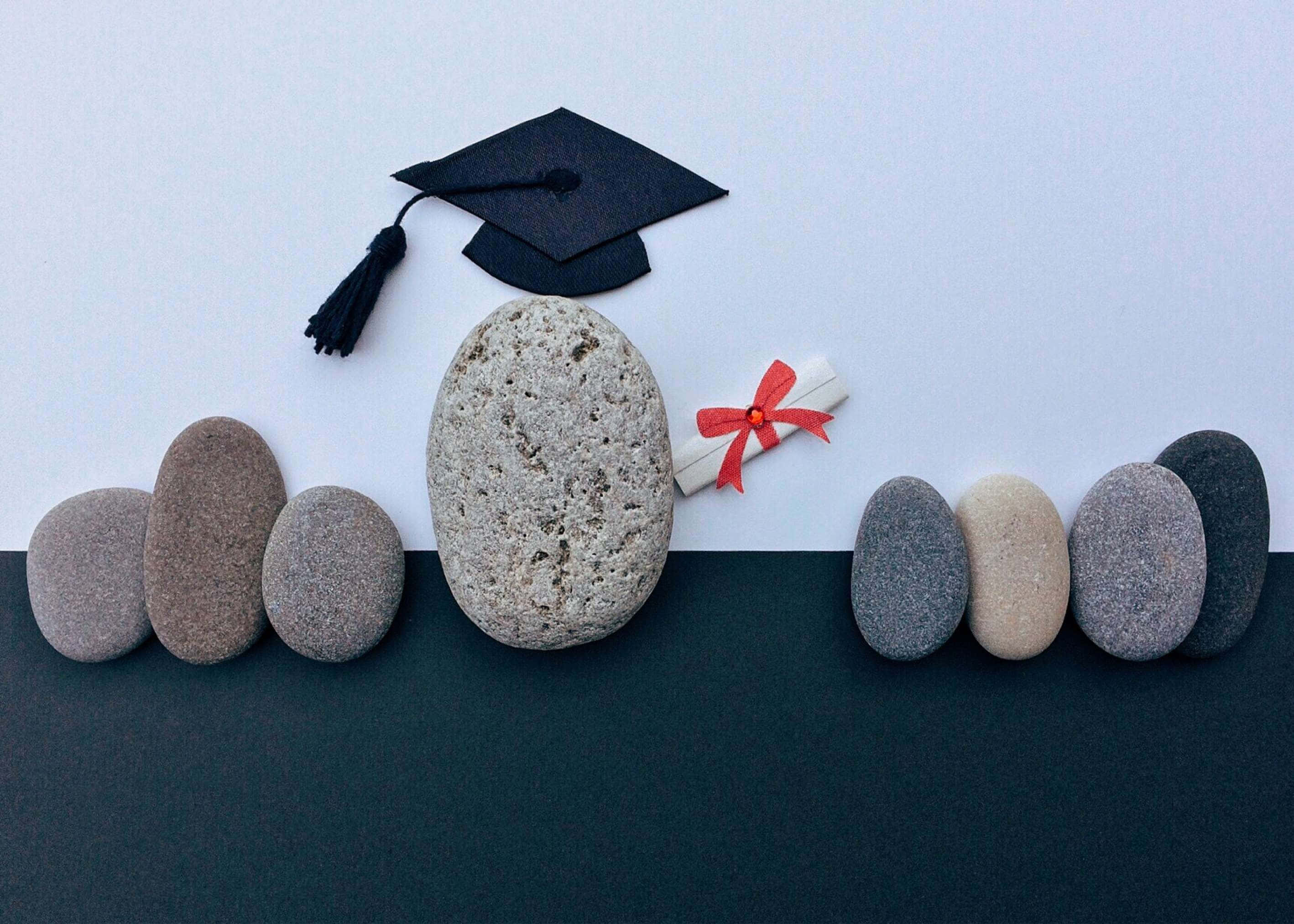 Graduation Ceremony 桌布 壁紙 — 畢業帽和文憑放在中央的石頭上，周圍是光滑的鵝卵石，背景為雙色調。