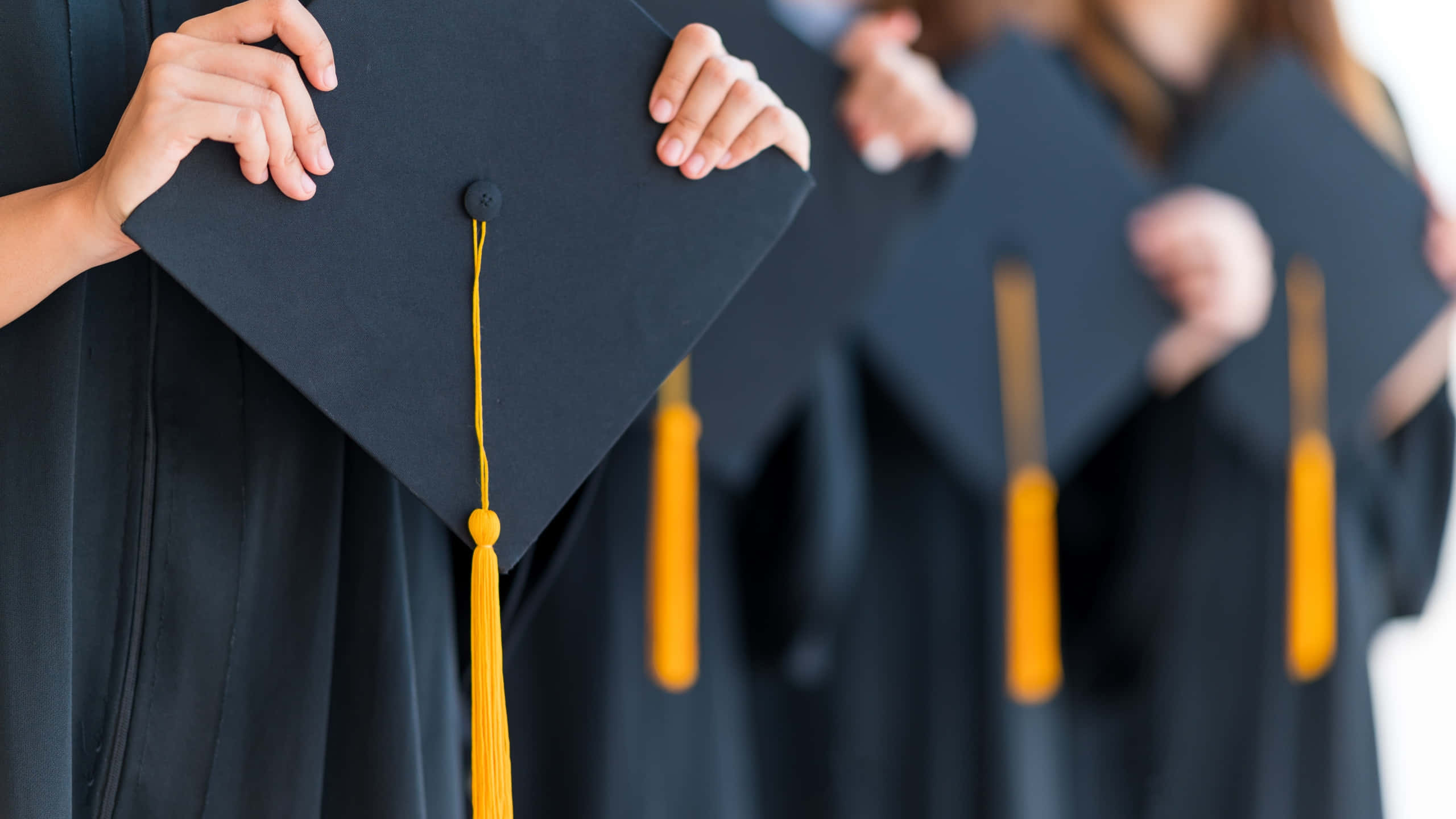 Graduation Ceremony 桌布 壁紙 — 畢業生手持黑色學位帽，帽上帶有亮黃色流蘇，背景為柔和白色的特寫。