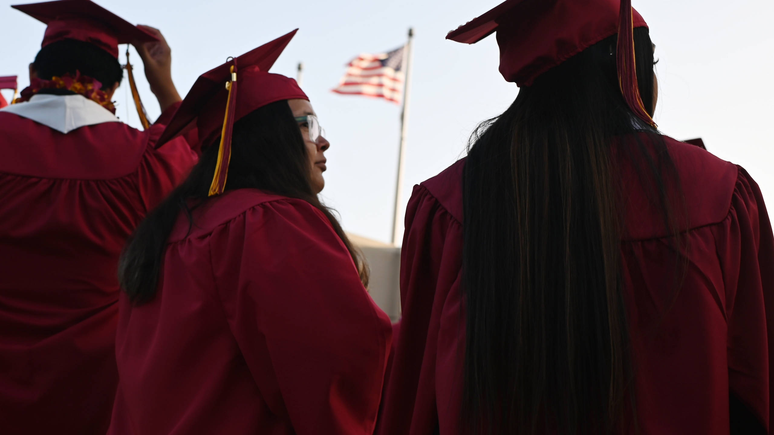 Graduation 桌布 壁紙 — 畢業生身穿栗色學位袍和學士帽，在日間典禮中莊嚴地站立，背景中美國國旗飄揚。
