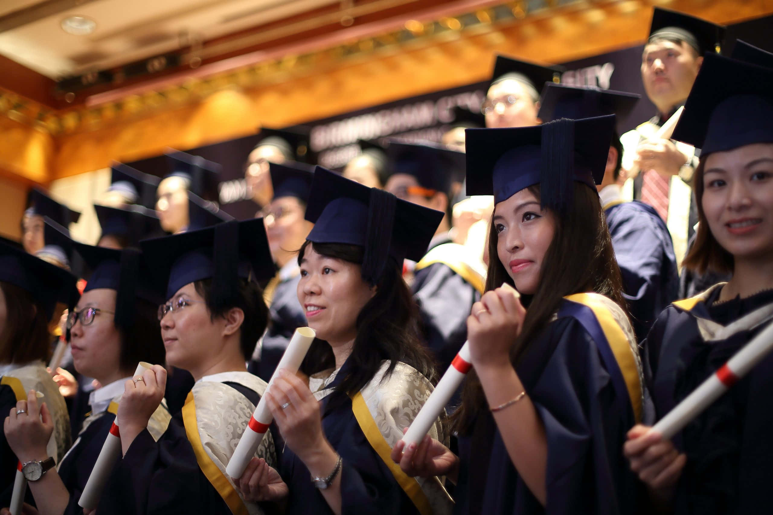 College Graduation 桌布 壁紙 — 畢業生身穿深色學士袍和四方帽，在溫暖燈光的禮堂中自豪地微笑著手持文憑。