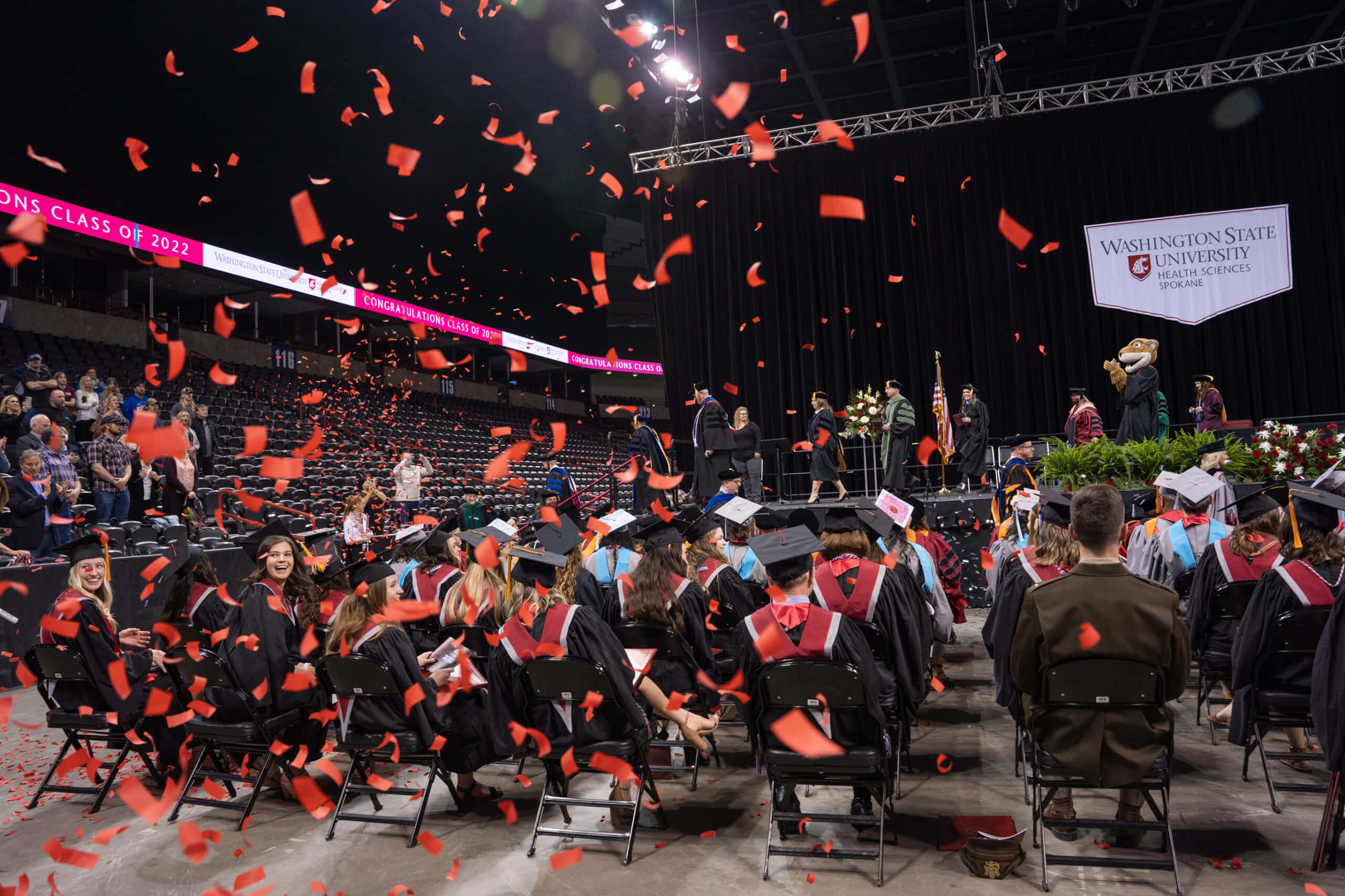 Nurses Graduation 桌布 壁紙 — 畢業生身穿黑色學位帽和袍，在擠滿人的場館內慶祝，紅色紙屑如雨般灑落，進行大學畢業典禮。