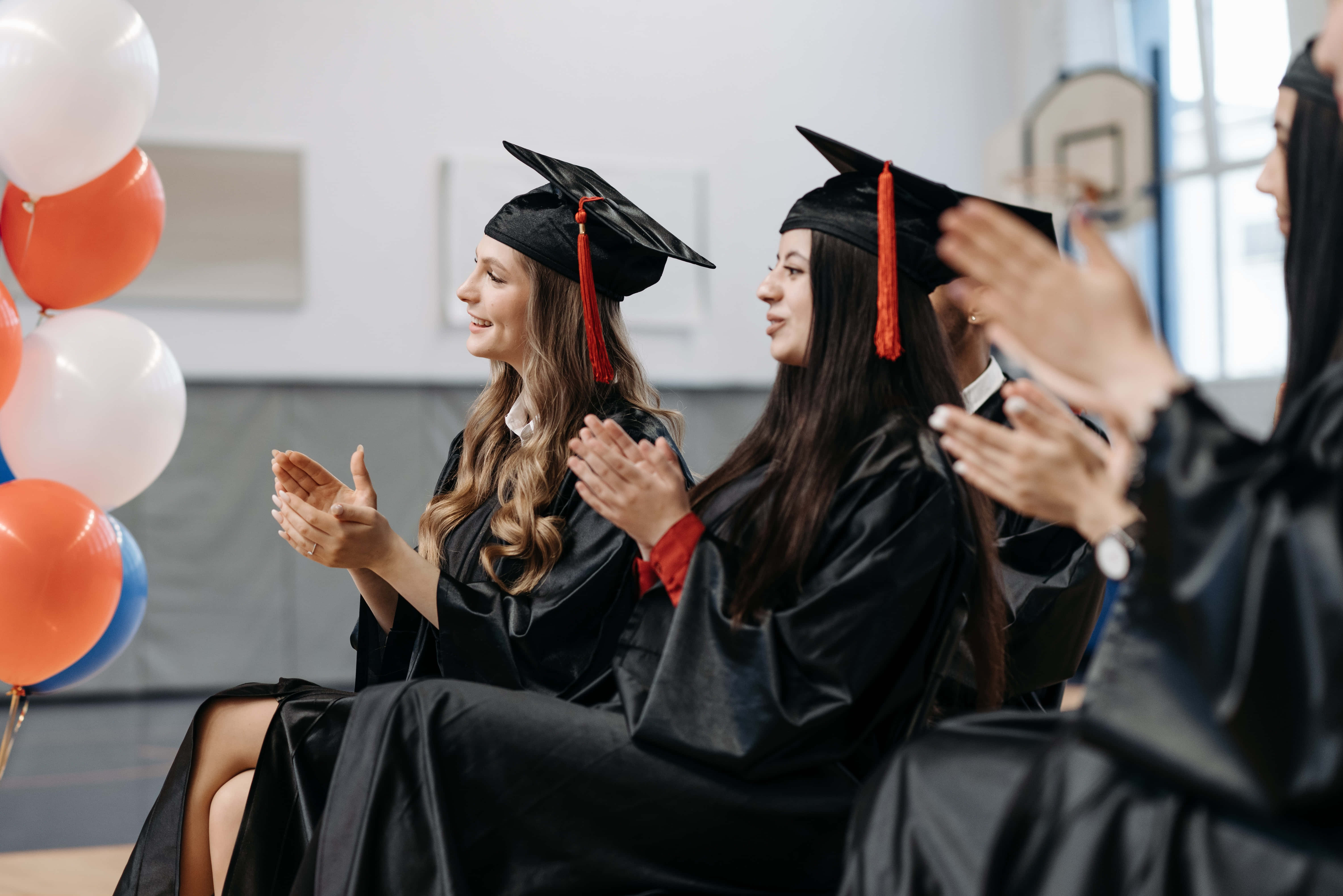 Graduation Ceremony 桌布 壁紙 — 畢業生身穿黑色學士帽和畢業袍，在明亮的體育館內，於色彩繽紛的氣球旁開心地鼓掌。
