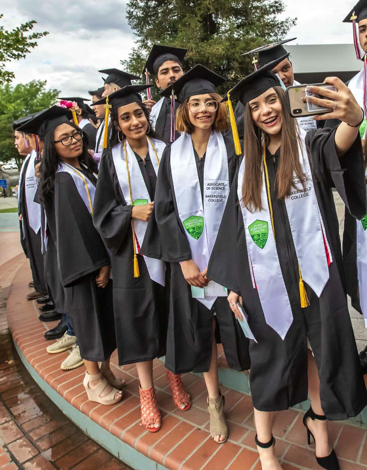 Graduation 桌布 壁紙 — 畢業生身穿黑色學士帽和袍，在陰天下磚砌小徑上，一人自拍，眾人燦爛微笑。
