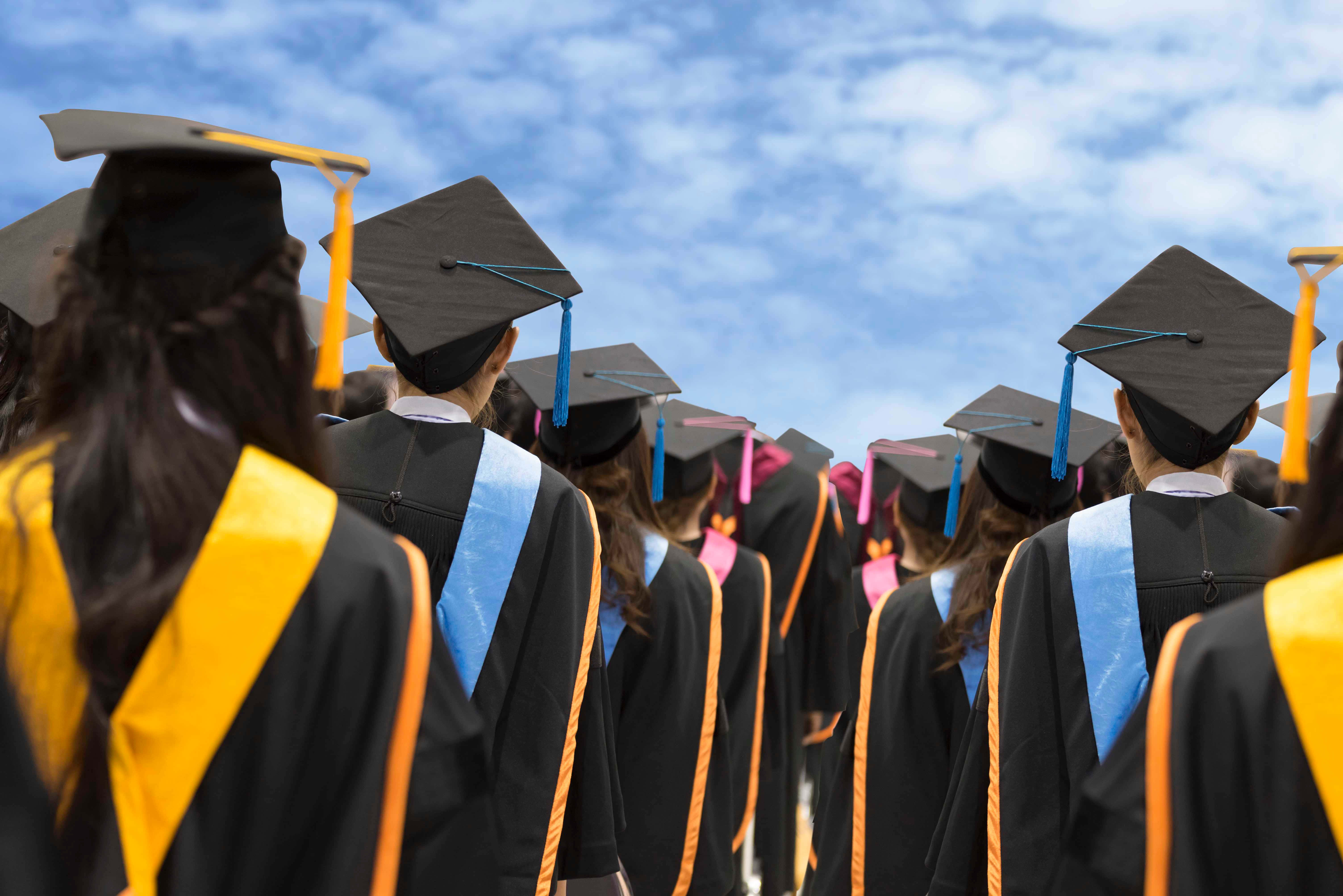 College Graduation 桌布 壁紙 — 畢業生身穿黑色學士帽和袍，戴著彩色流蘇，在晴朗藍天白雲下行走的背影。