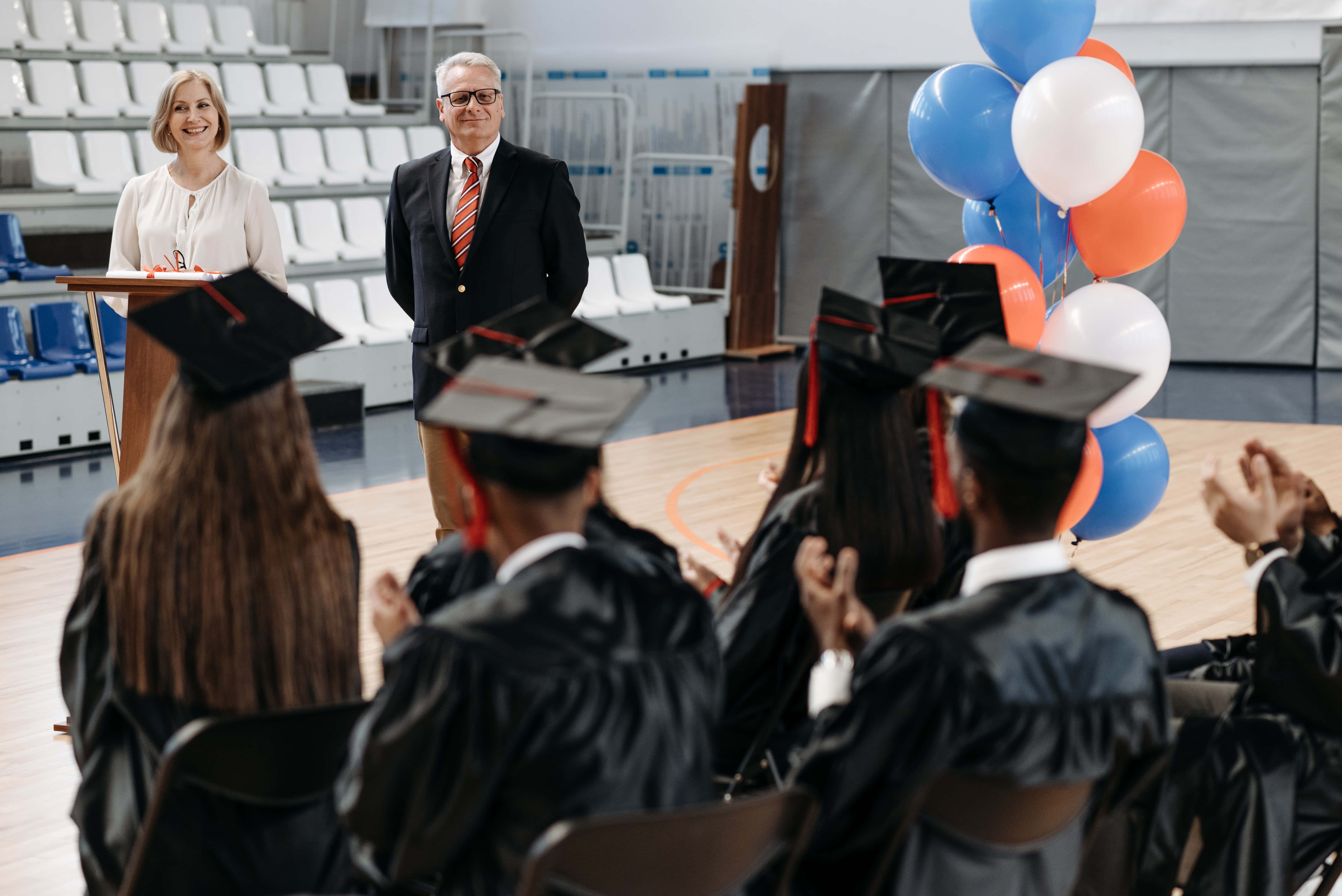 Graduation Ceremony 桌布 壁紙 — 畢業生身穿黑色學士帽和袍鼓掌，教職員站在以藍色、橙色和白色氣球裝飾的體育館講台上。