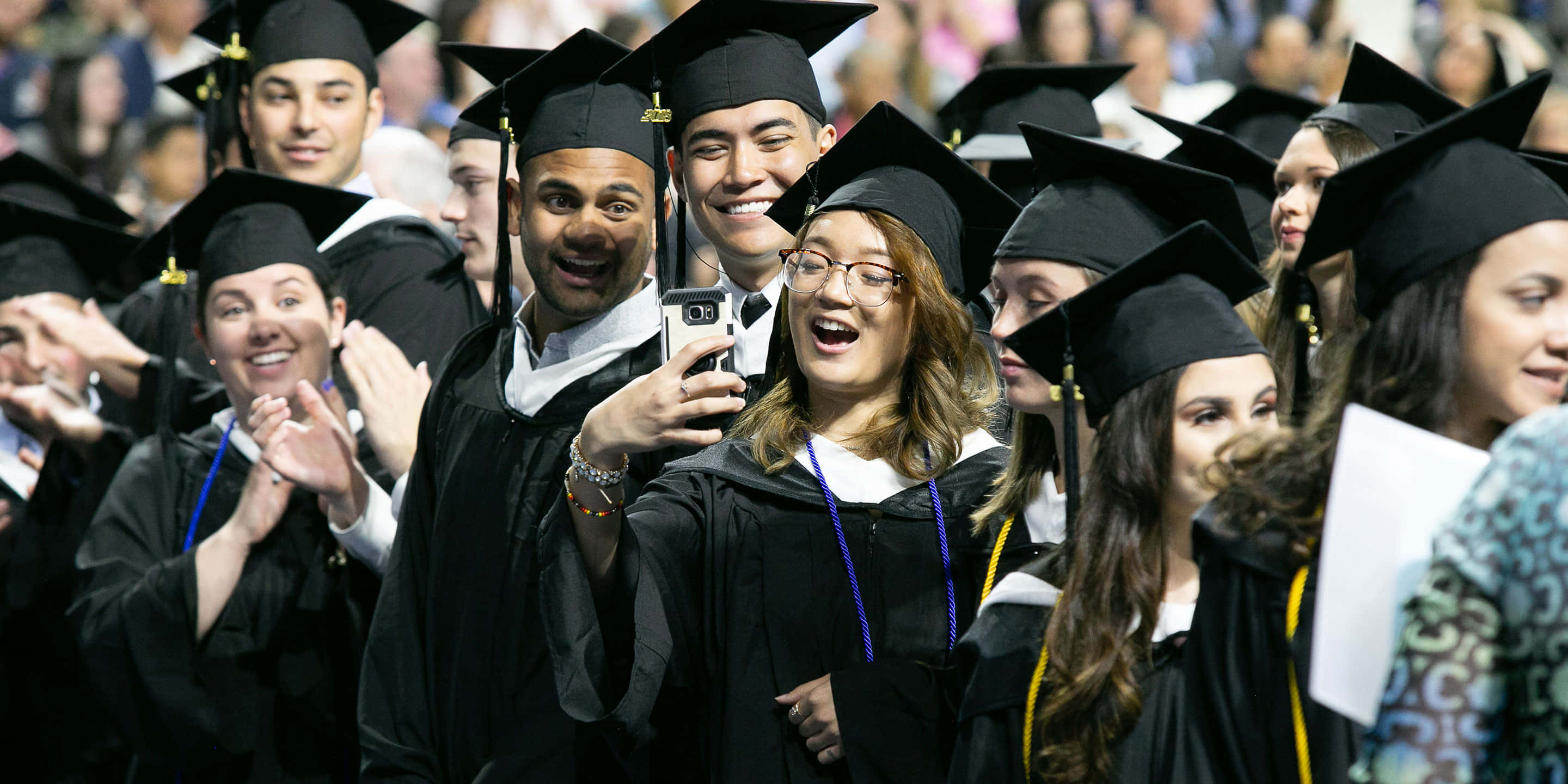 Graduation 桌布 壁紙 — 畢業生身穿黑色學士袍和學士帽，在擠滿人的畢業典禮上開心地自拍。