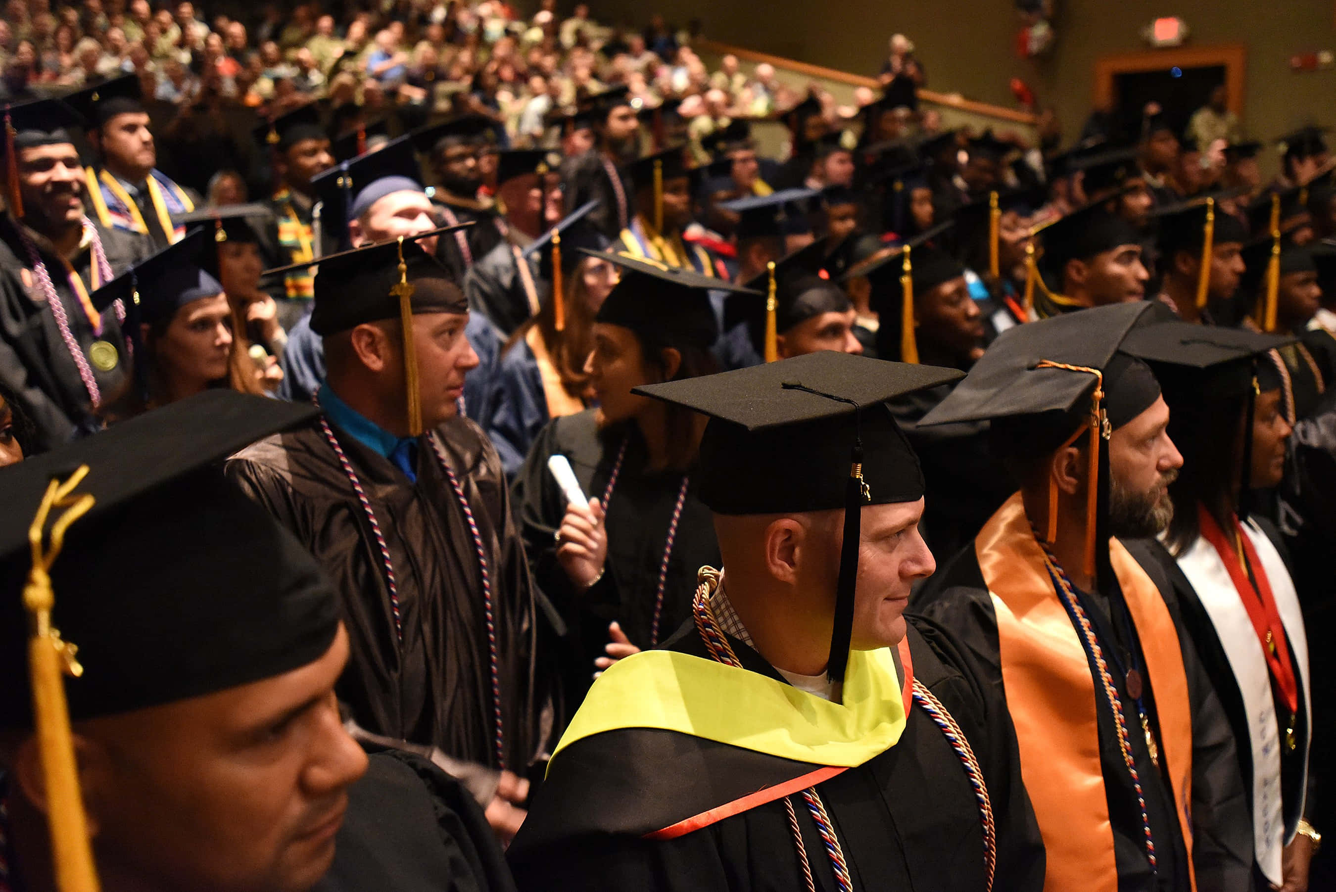 Graduation 桌布 壁紙 — 畢業生身穿黑色學士袍和方帽，在溫暖的室內燈光下，於擁擠的畢業典禮上排成行列。