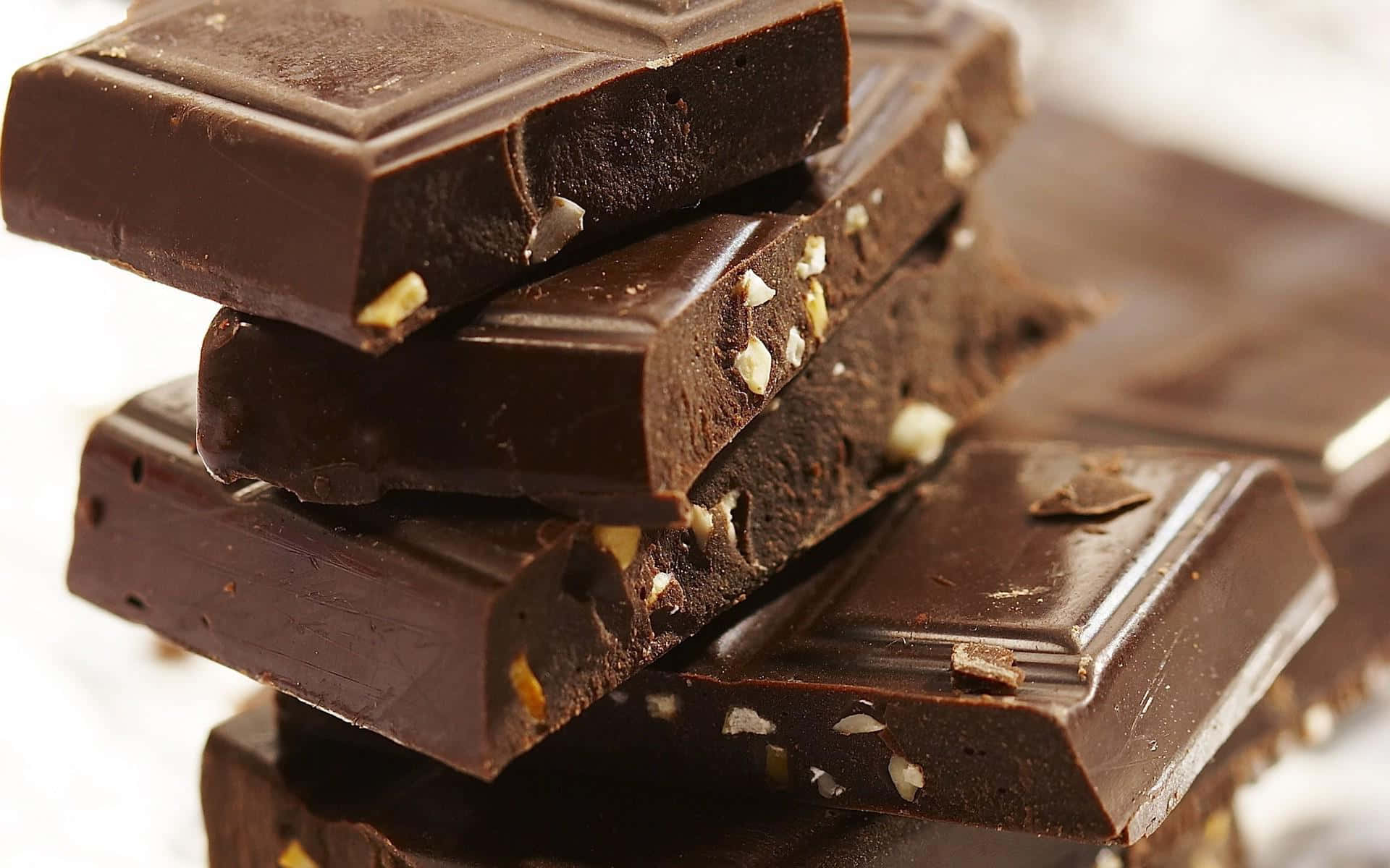 Brown Chocolate 桌布 壁紙 — 疊起的黑巧克力條帶有可見的堅果碎塊，在柔和光線下於模糊的白色背景上閃閃發光。