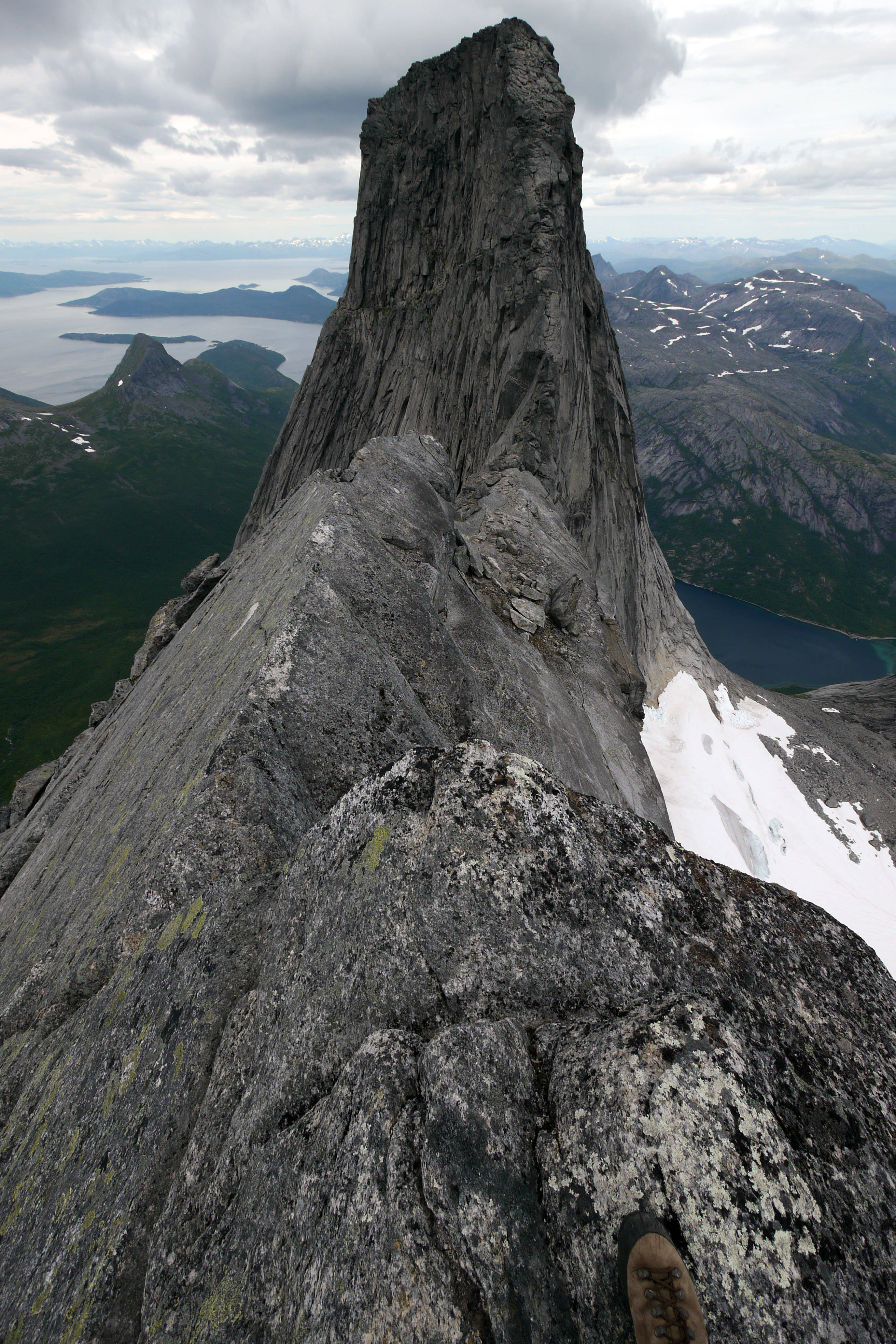 Norway 桌布 壁紙 — 登山者的靴子擱在鋸齒狀的灰色岩石山峰上，俯瞰著廣闊的峽灣和白雪覆蓋的山脈，天空多雲。