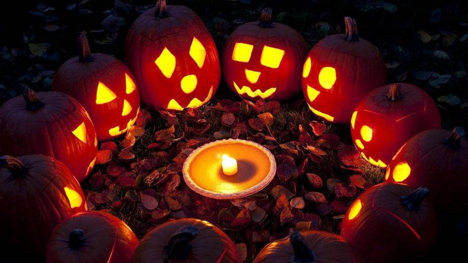 Halloween Phone 桌布 壁紙 — 發光的南瓜燈籠帶有雕刻的臉孔，圍繞著一根點燃的蠟燭，放在暗淡光線下的秋葉堆上。