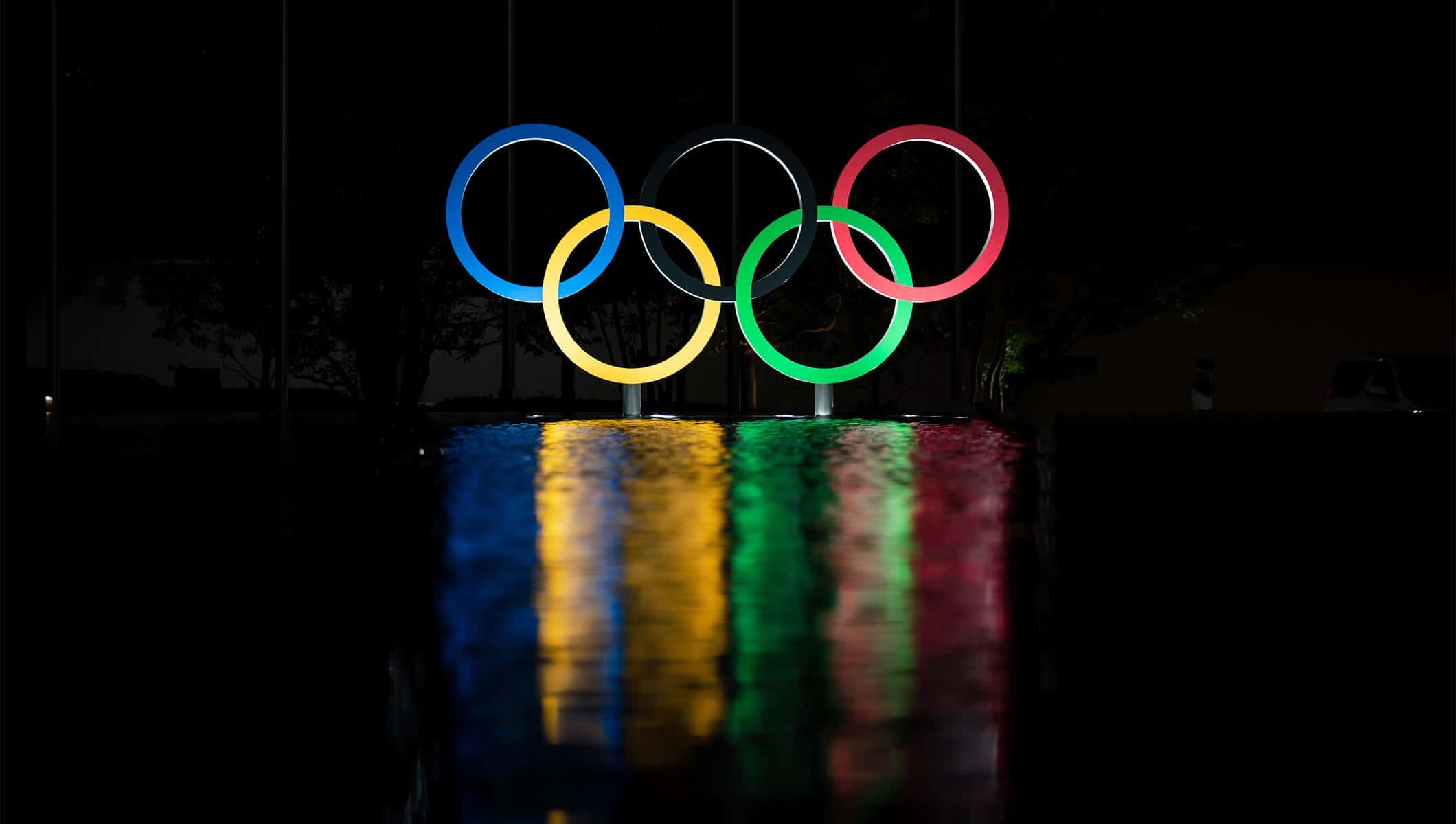 Olympics 桌布 壁紙 — 發光的奧運五環在夜晚閃耀，下方濕漉漉的人行道上倒映著生動的反射光芒。
