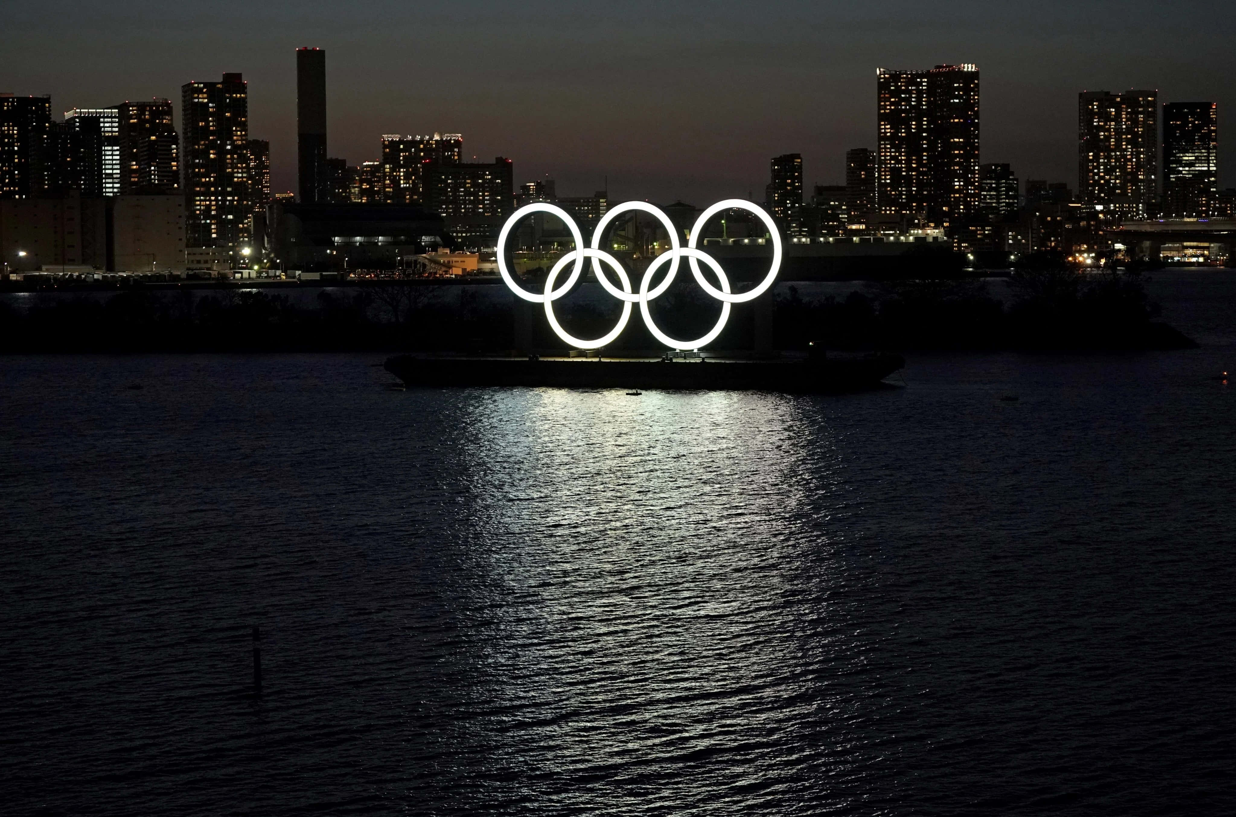 Olympics 桌布 壁紙 — 發光的奧運五環在黃昏時漂浮在水面上，背後的城市天際線閃爍著光芒。