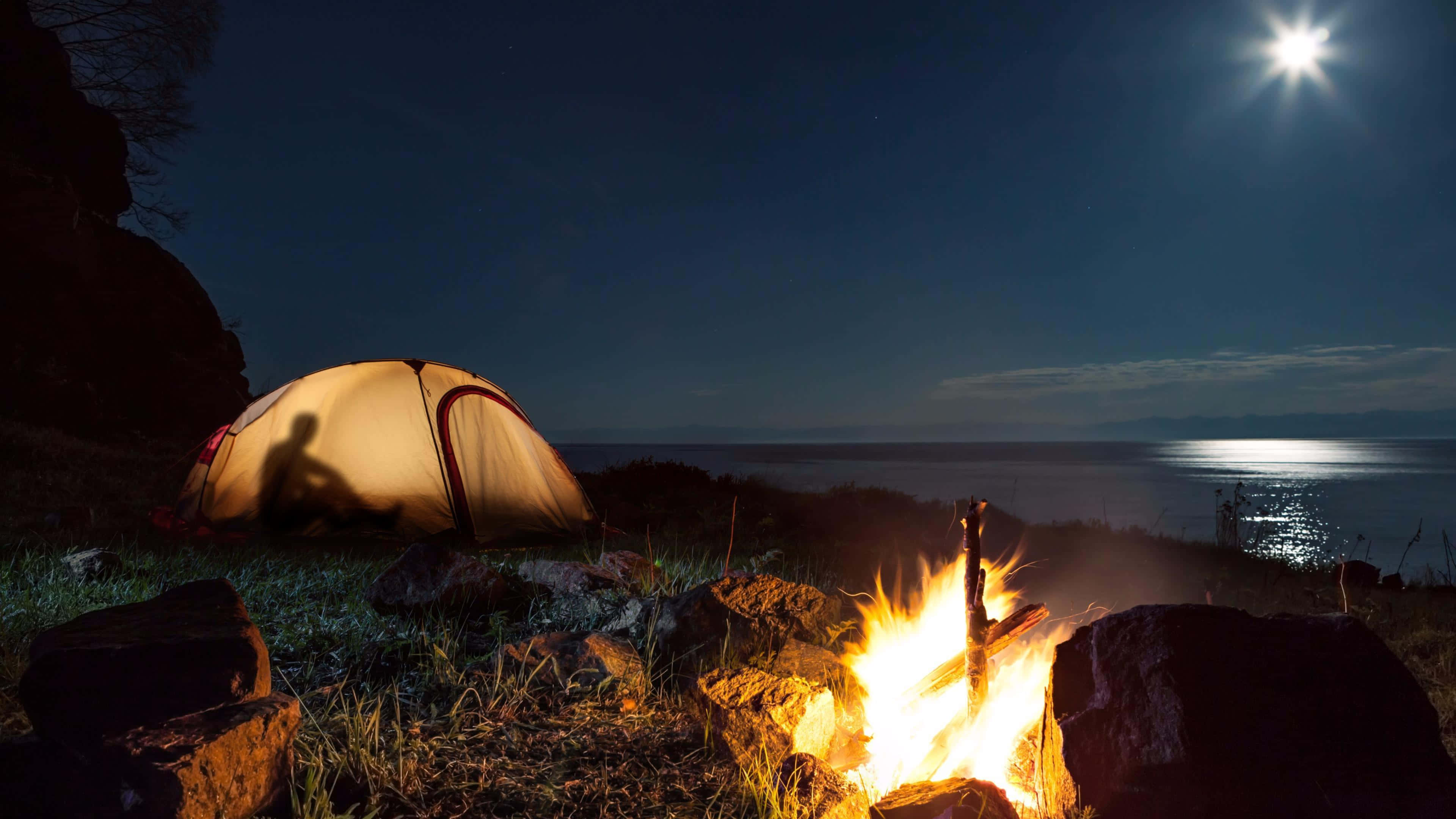 Beach Camping 桌布 壁紙 — 發光的帳篷和營火照亮了岩石海岸，夜空下平靜的海面上掛著明亮的月亮。