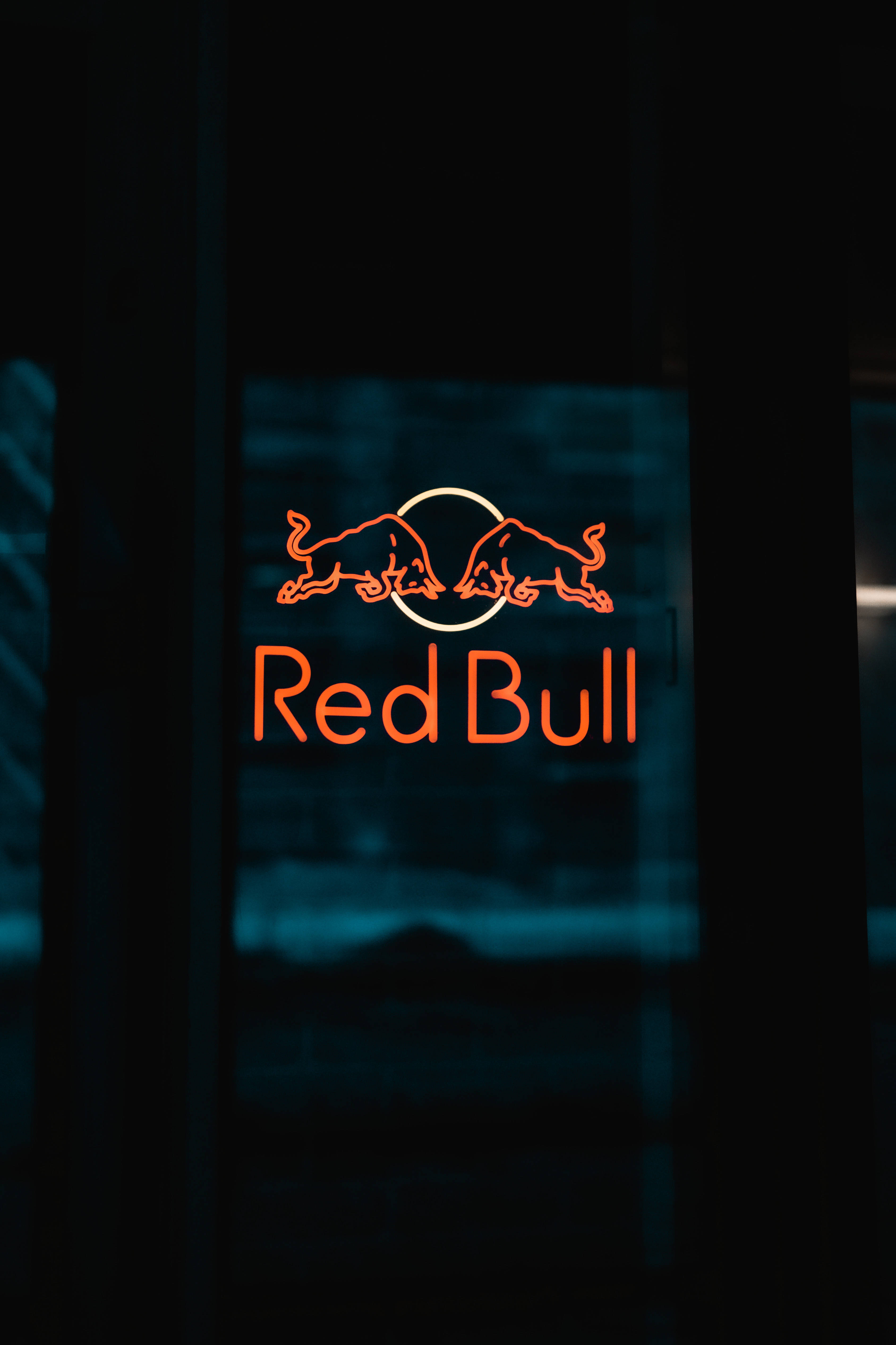 Red Bull 桌布 壁紙 — 發光的橙色紅牛標誌霓虹燈牌在帶有藍色反射的深色玻璃門上明亮地閃耀。