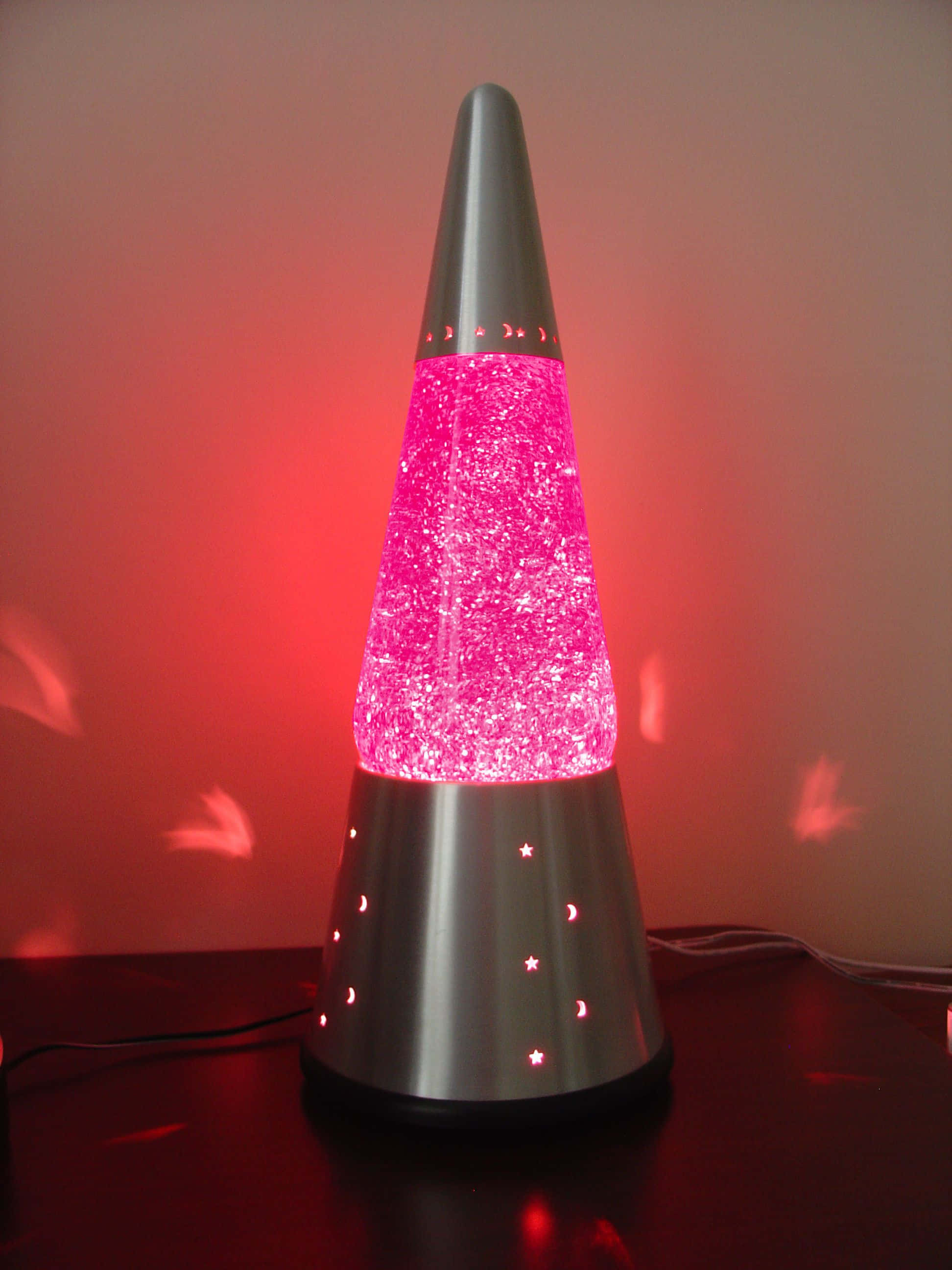 lava lamp 桌布 壁紙 — 發光的粉紅色熔岩燈，帶有銀色圓錐底座和星星月亮鏤空，在牆上投射出紅光。