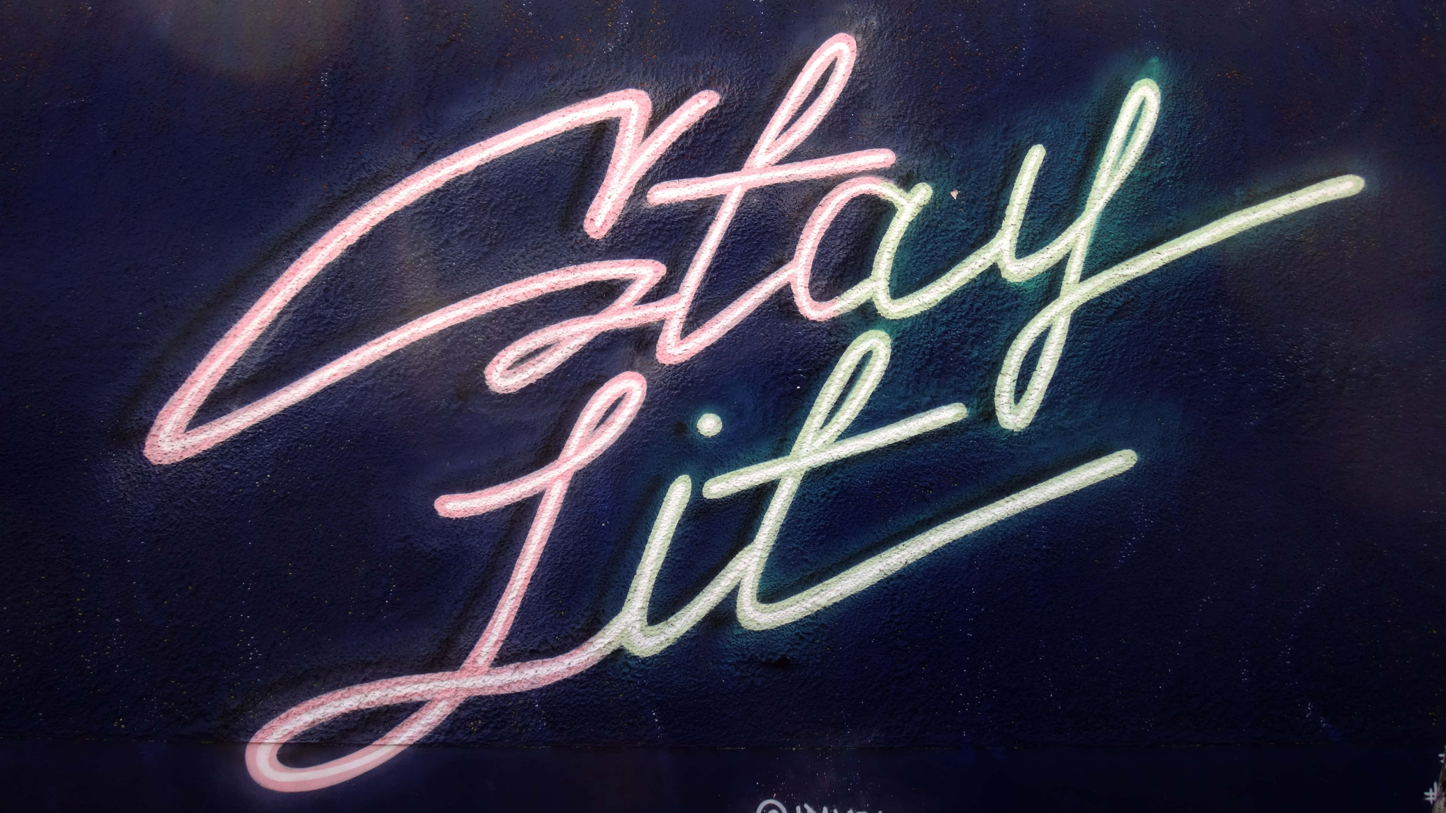 Lit iPhone 桌布 壁紙 — 發光的霓虹燈字體在深色紋理牆壁上顯示「Stay Lit」，帶有粉紅色和綠色色調。