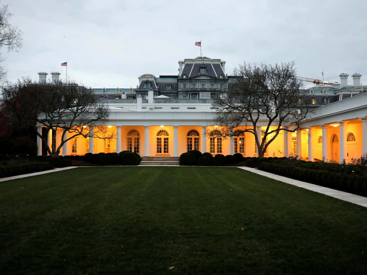 White House Rose Garden 桌布 壁紙 — 白宮在暮色天空下溫暖地發光，光禿的樹木襯托著其宏偉的柱子和修剪整齊的草坪。
