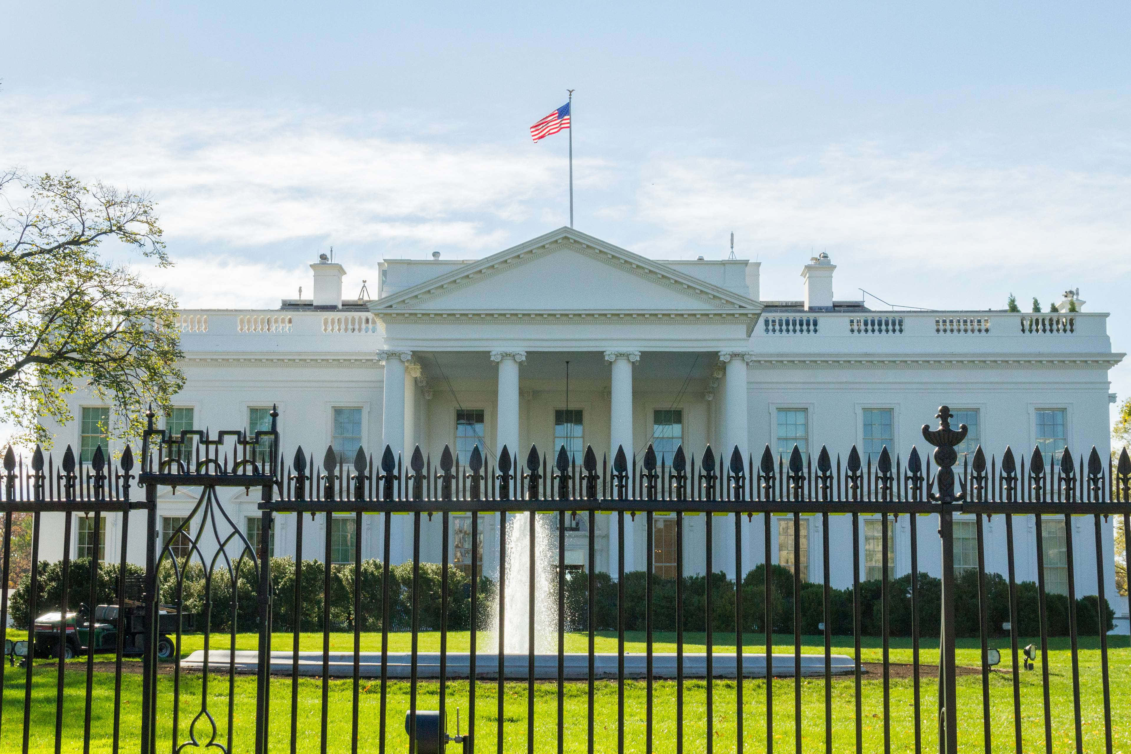 white house 桌布 壁紙 — 白宮在黑色鐵欄杆後莊嚴矗立，上方有美國國旗在晴朗藍天下飄揚。