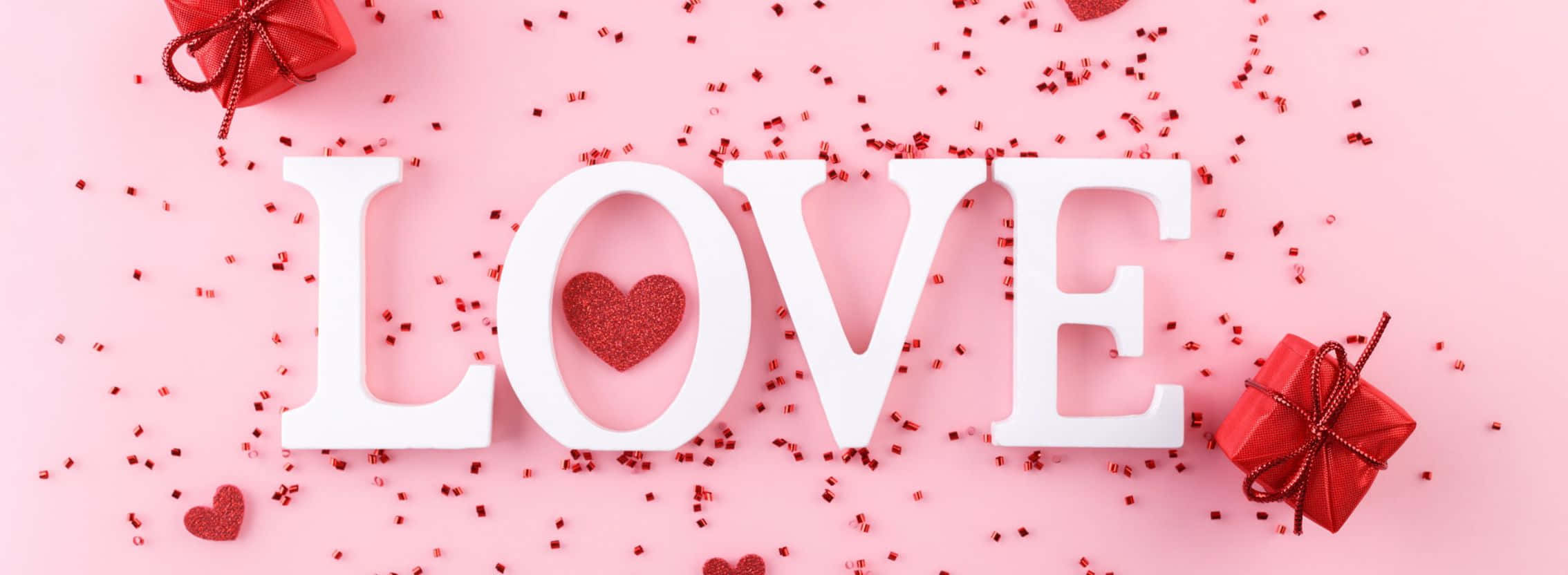 2268x832 桌布 壁紙 — 白色 'LOVE' 字母，中心有閃亮的心形，周圍是紅色五彩紙屑和禮物盒，背景為柔和的粉紅色。