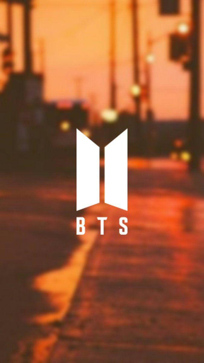 BTS標誌出現在黃昏時分模糊的濕漉街道上，帶有溫暖的橙色燈光。 — BTS標誌 壁紙