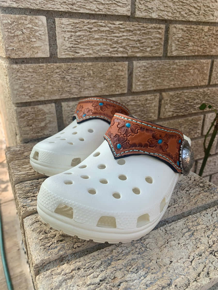 Crocs 桌布 壁紙 — 白色Crocs鞋，飾有華麗的棕色皮革帶和綠松石飾釘，擱在帶紋理的石階上，背景是磚牆。