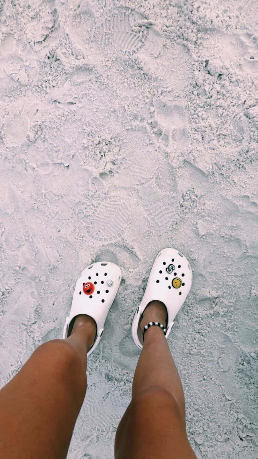 Crocs 桌布 壁紙 — 白色Crocs鞋配上色彩繽紛的吊飾，站立在質感沙地上，周圍可見腳印。