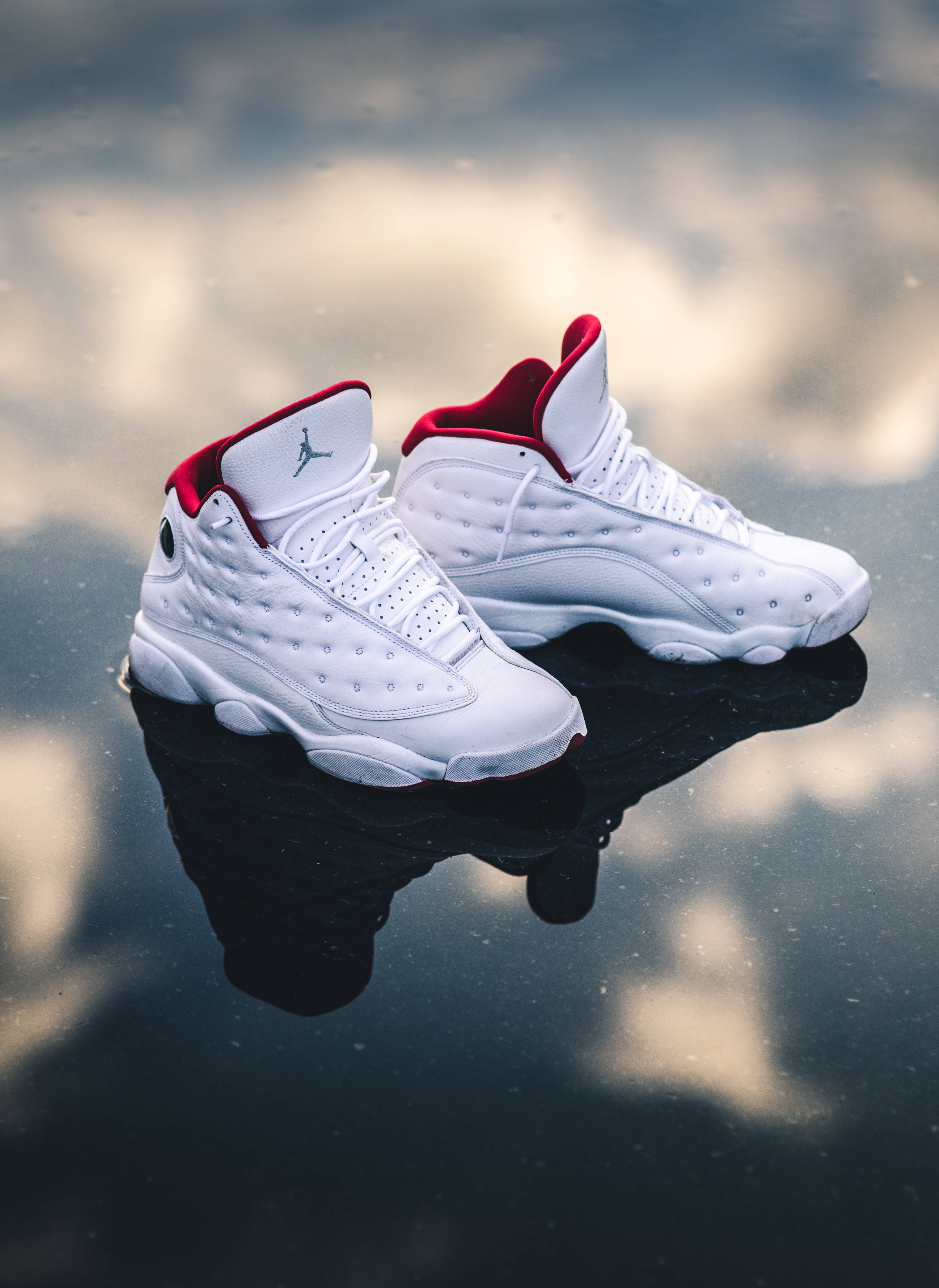 Air Jordan 桌布 壁紙 — 白色Jordan 13運動鞋帶有紅色點綴，放在潮濕的路面上，反射出多雲的天空。