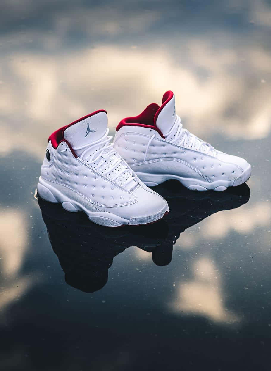 白色Jordan 13運動鞋帶有紅色點綴，置於反光的黑色表面上。 — Air Jordan 1 壁紙