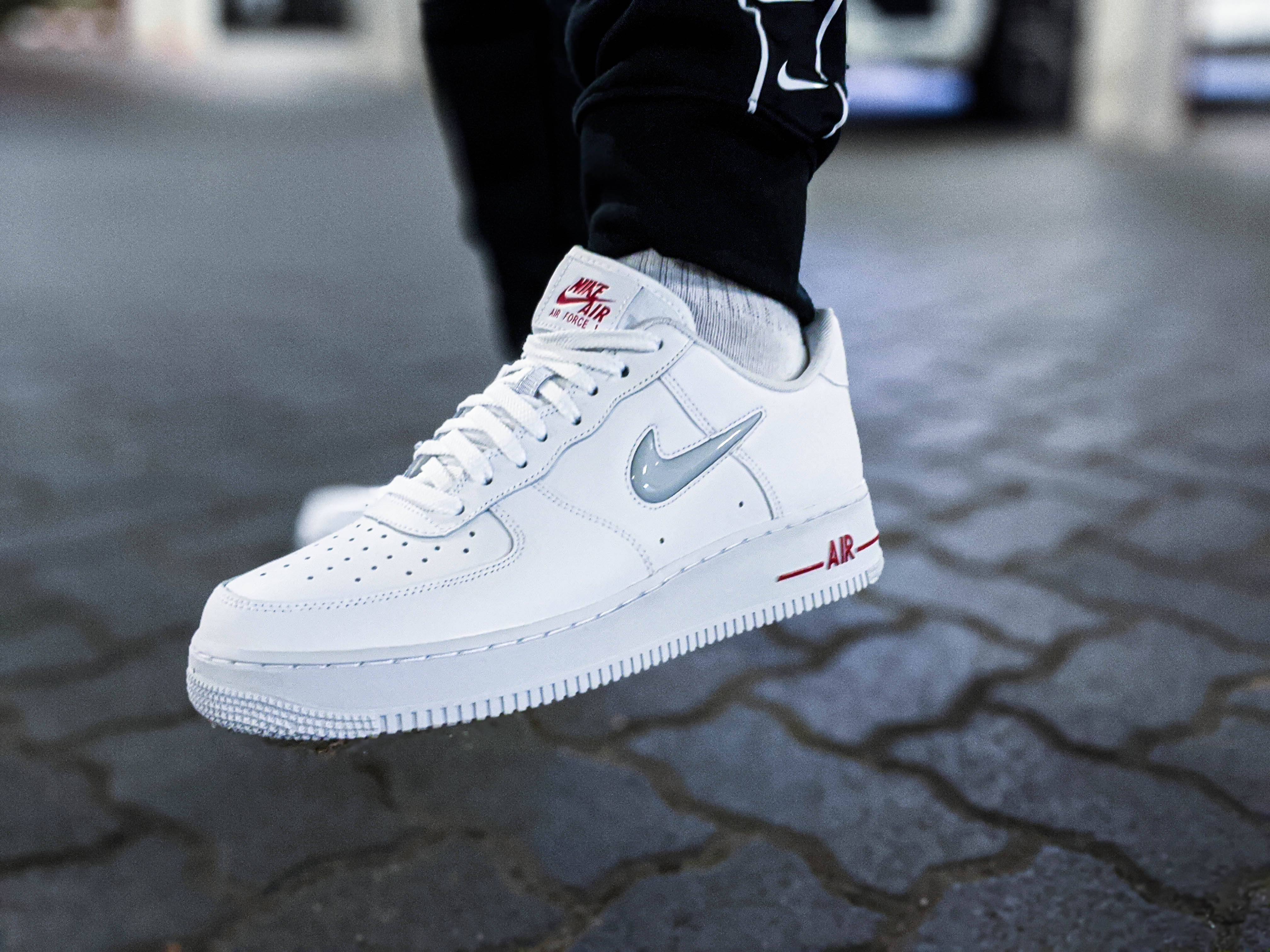 Shoe 桌布 壁紙 — 白色Nike Air Force 1運動鞋特寫，帶有紅色和銀色點綴，放在鵝卵石街道上。