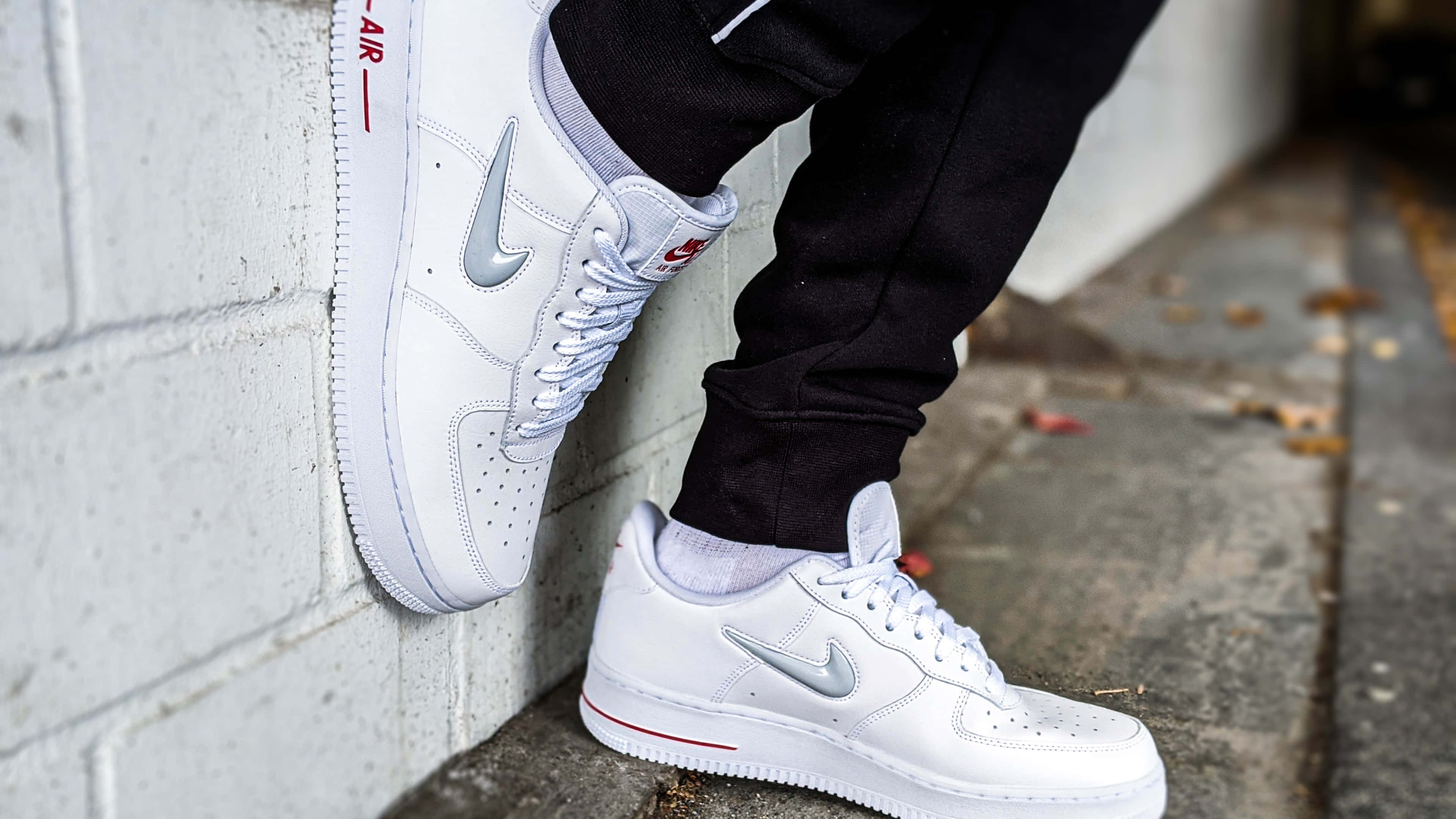 桌布 壁紙 — 白色Nike Air Force 1運動鞋特寫，帶有銀色swoosh和紅色點綴，由靠著混凝土牆的人穿著。