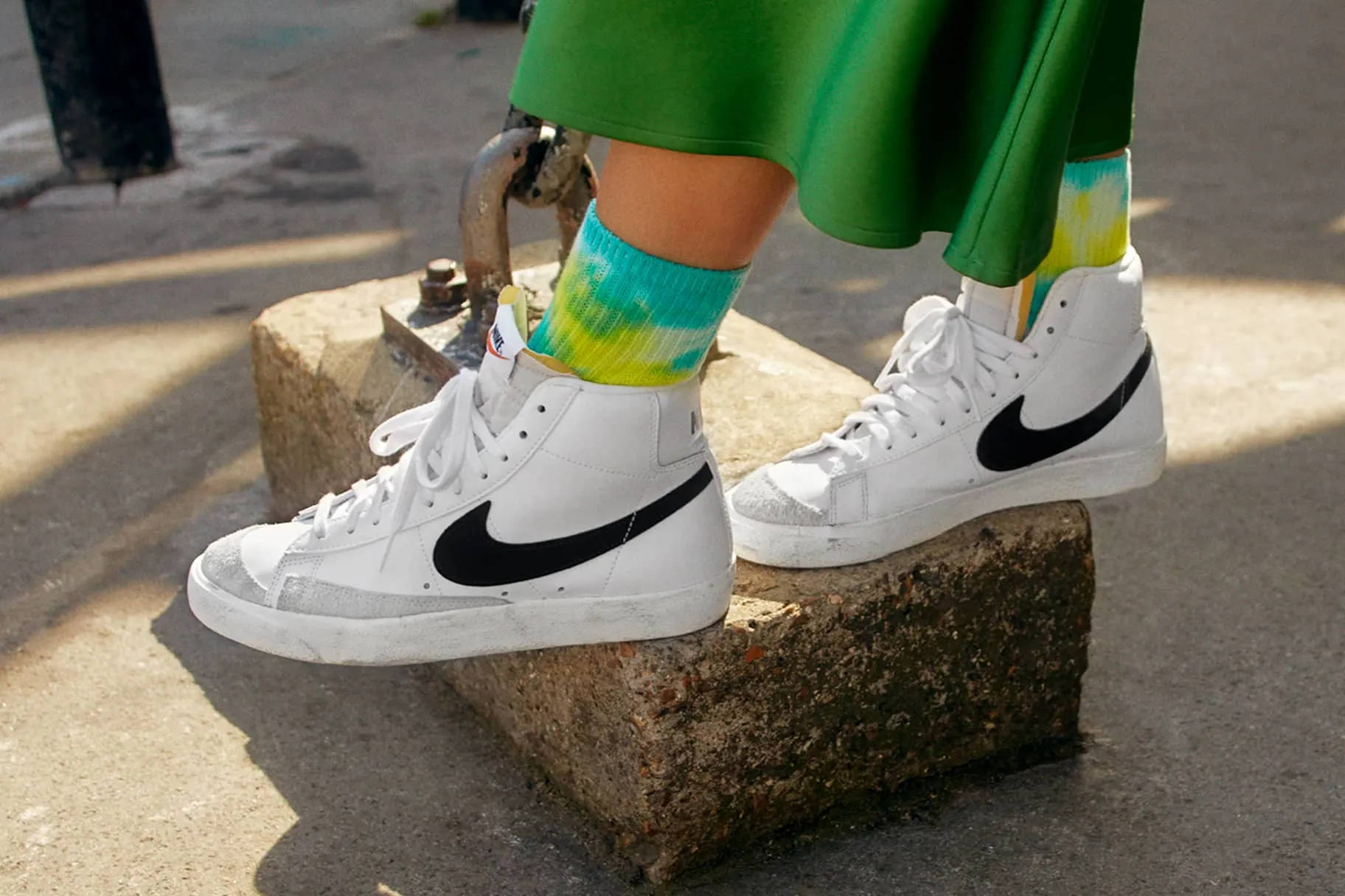 Nike Shoes 桌布 壁紙 — 白色Nike Blazer運動鞋搭配黑色swoosh，穿著紮染襪子和綠色裙子，在明亮陽光下的混凝土塊上特寫。