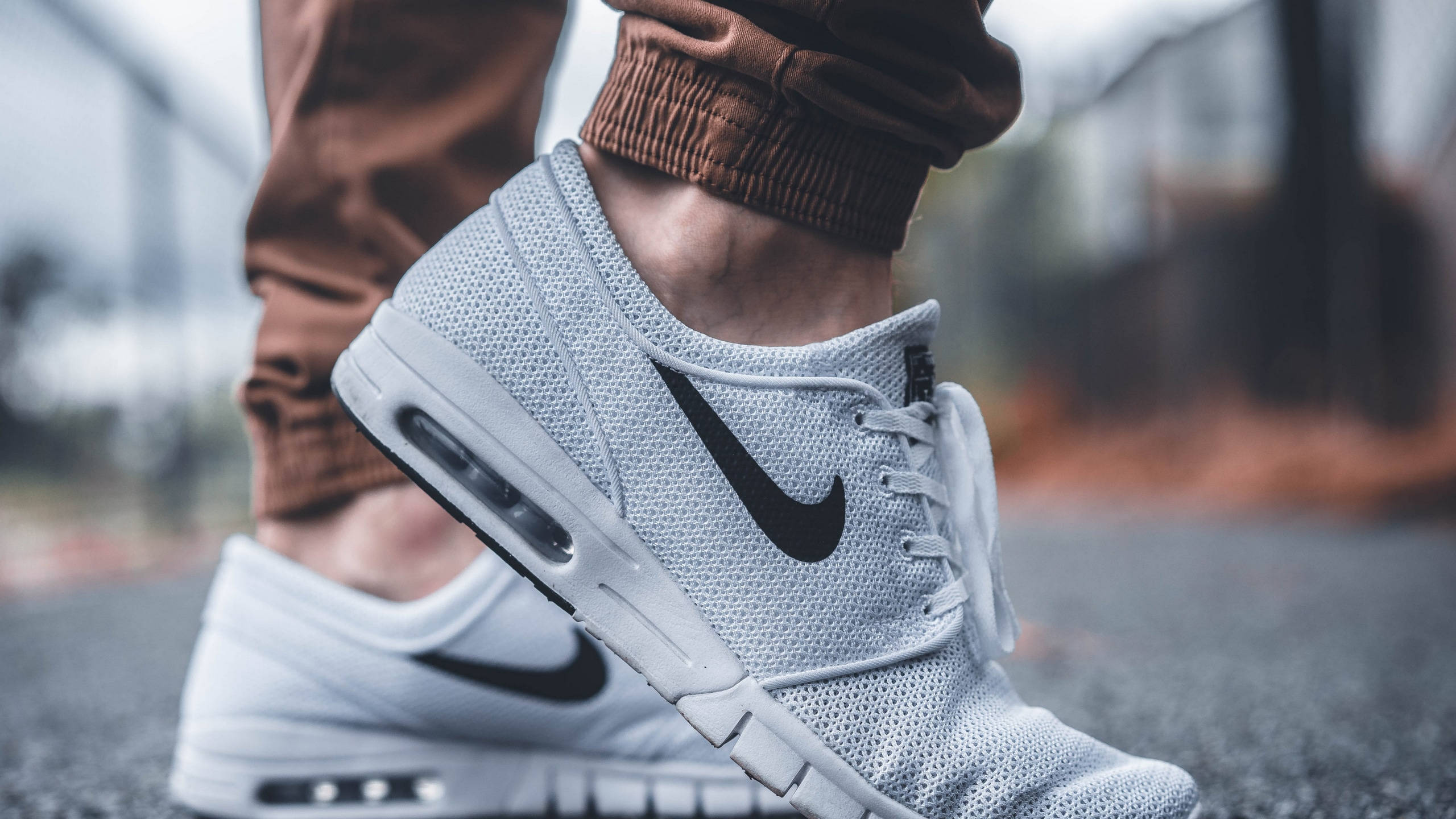 Nike Shoes 桌布 壁紙 — 白色Nike運動鞋特寫，帶有黑色swoosh標誌，由穿著棕色慢跑褲的人穿著在瀝青路面上。