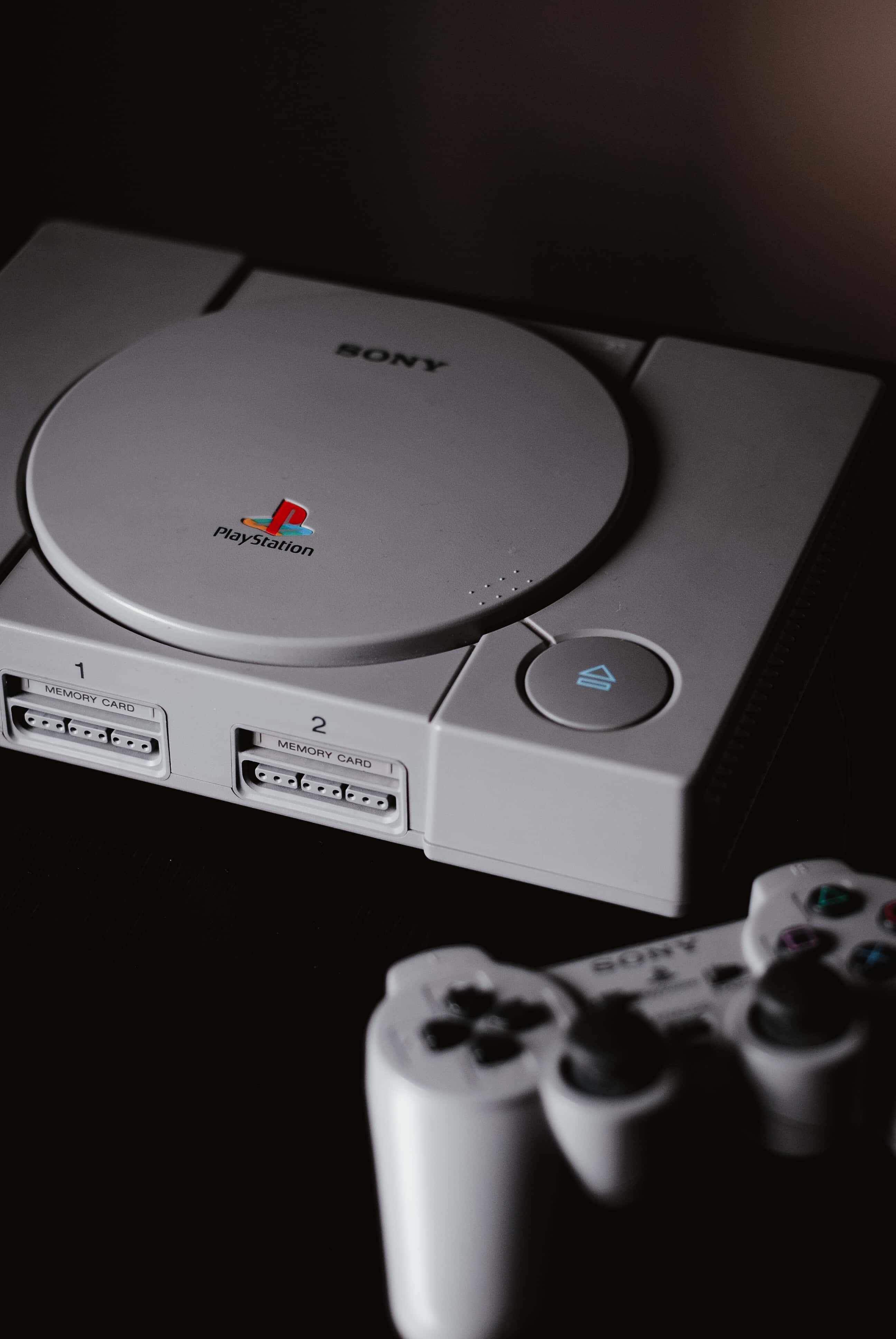 Gaming Consoles 桌布 壁紙 — 白色Sony PlayStation 1主機特寫，前景有控制器，背景為深色。