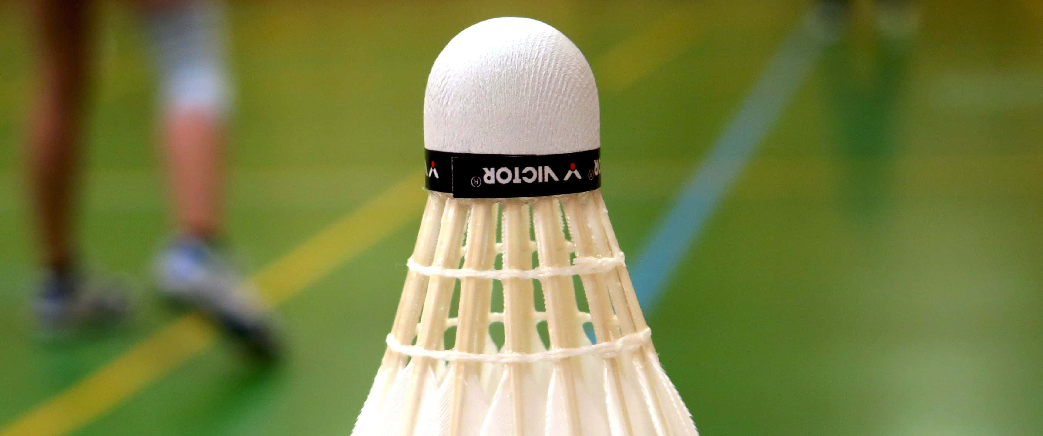 3440x1440p Badminton 桌布 壁紙 — 白色Victor羽毛球在綠色球場上的特寫，背景有模糊的球員腿部。