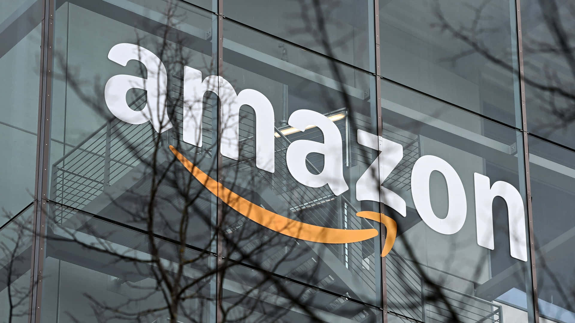 亞馬遜 桌布 壁紙 — 白色和橙色的Amazon標誌在玻璃外牆上，前景有光禿禿的樹枝剪影。