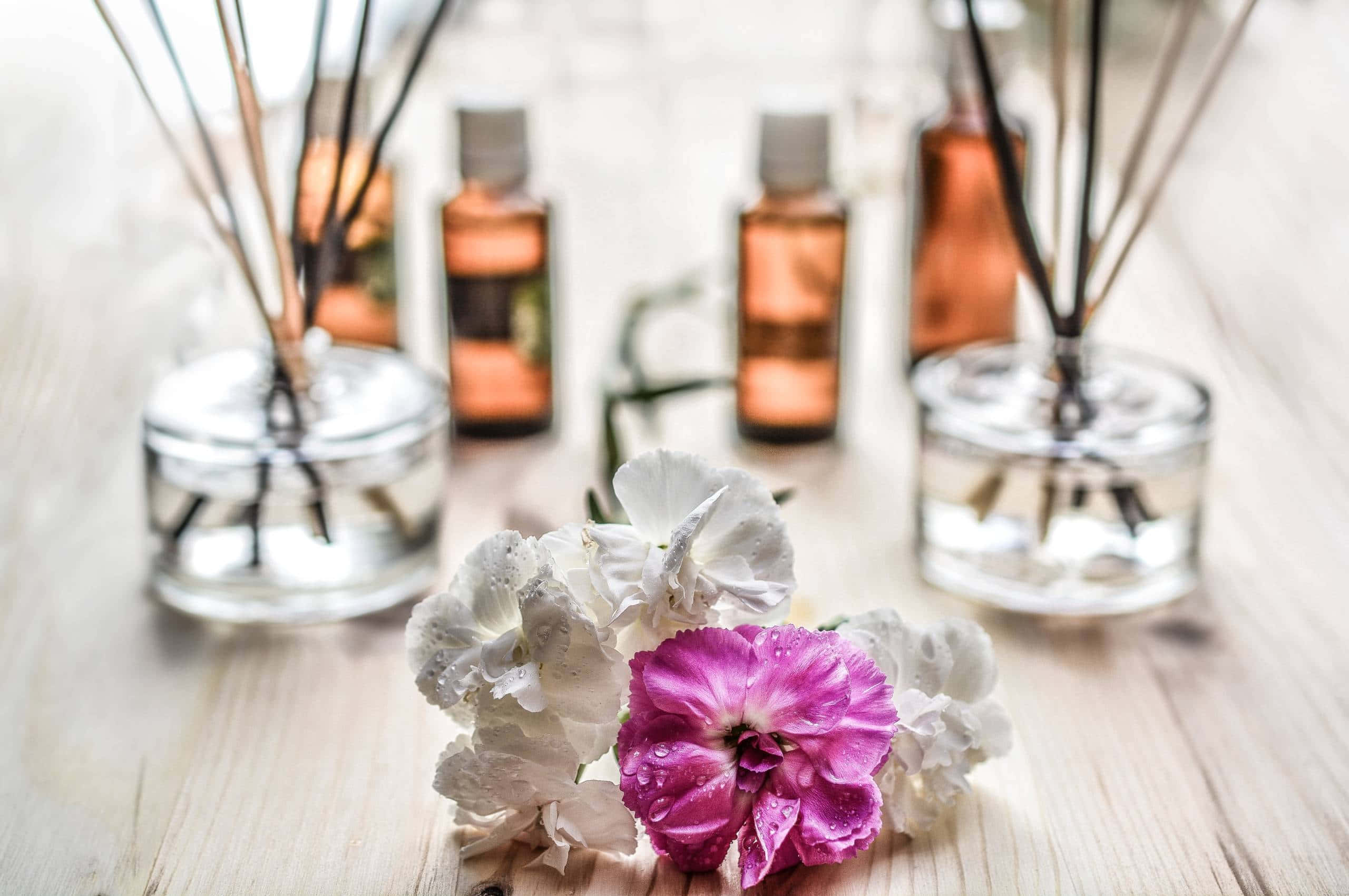Aromatherapy 桌布 壁紙 — 白色和粉紅色的康乃馨帶著露珠，放在木桌上，旁邊有玻璃擴香器和琥珀色瓶子。