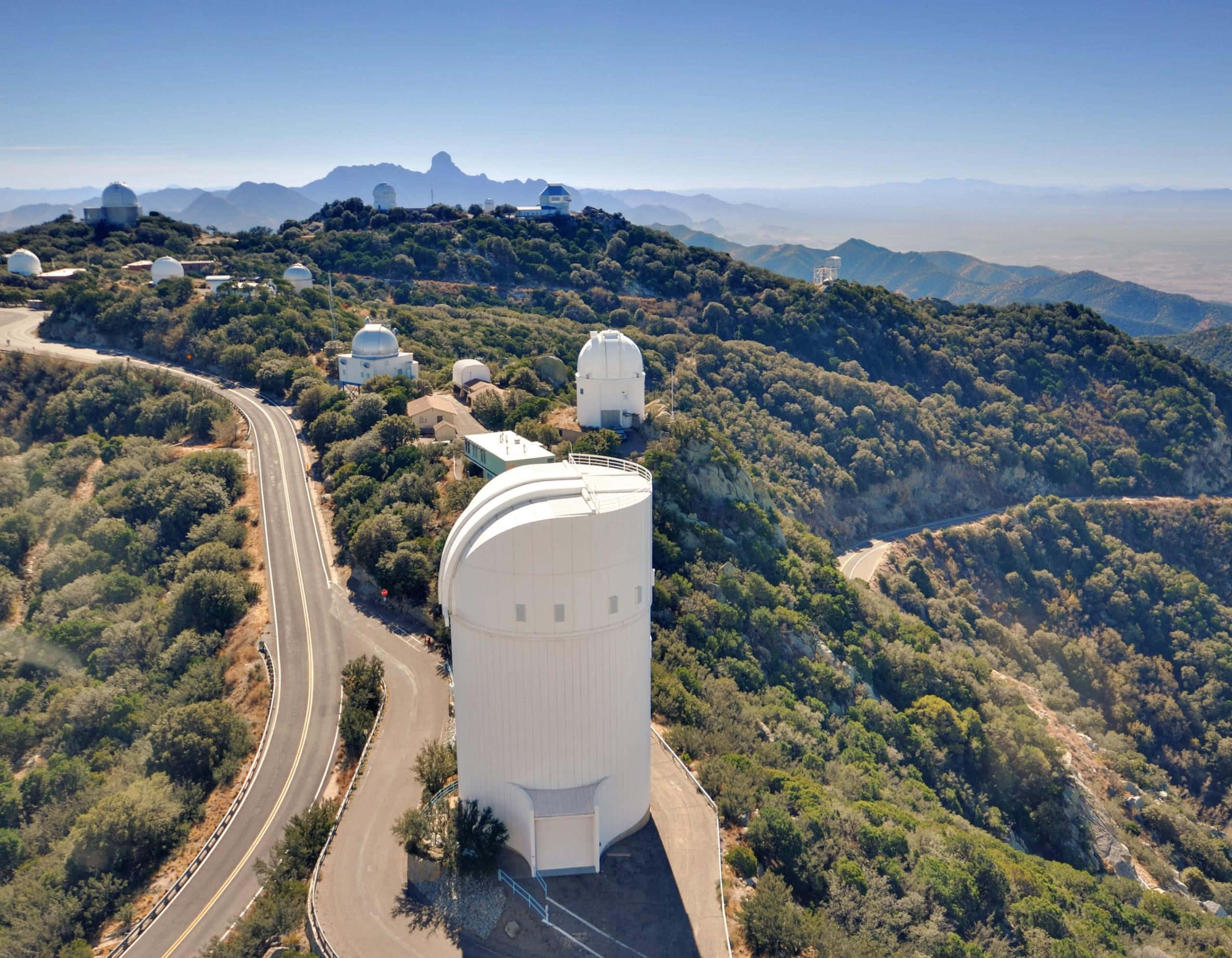 Space Observation 桌布 壁紙 — 白色天文台圓頂坐落在綠色山丘之間，蜿蜒的道路在晴朗藍天下延伸的鳥瞰圖。