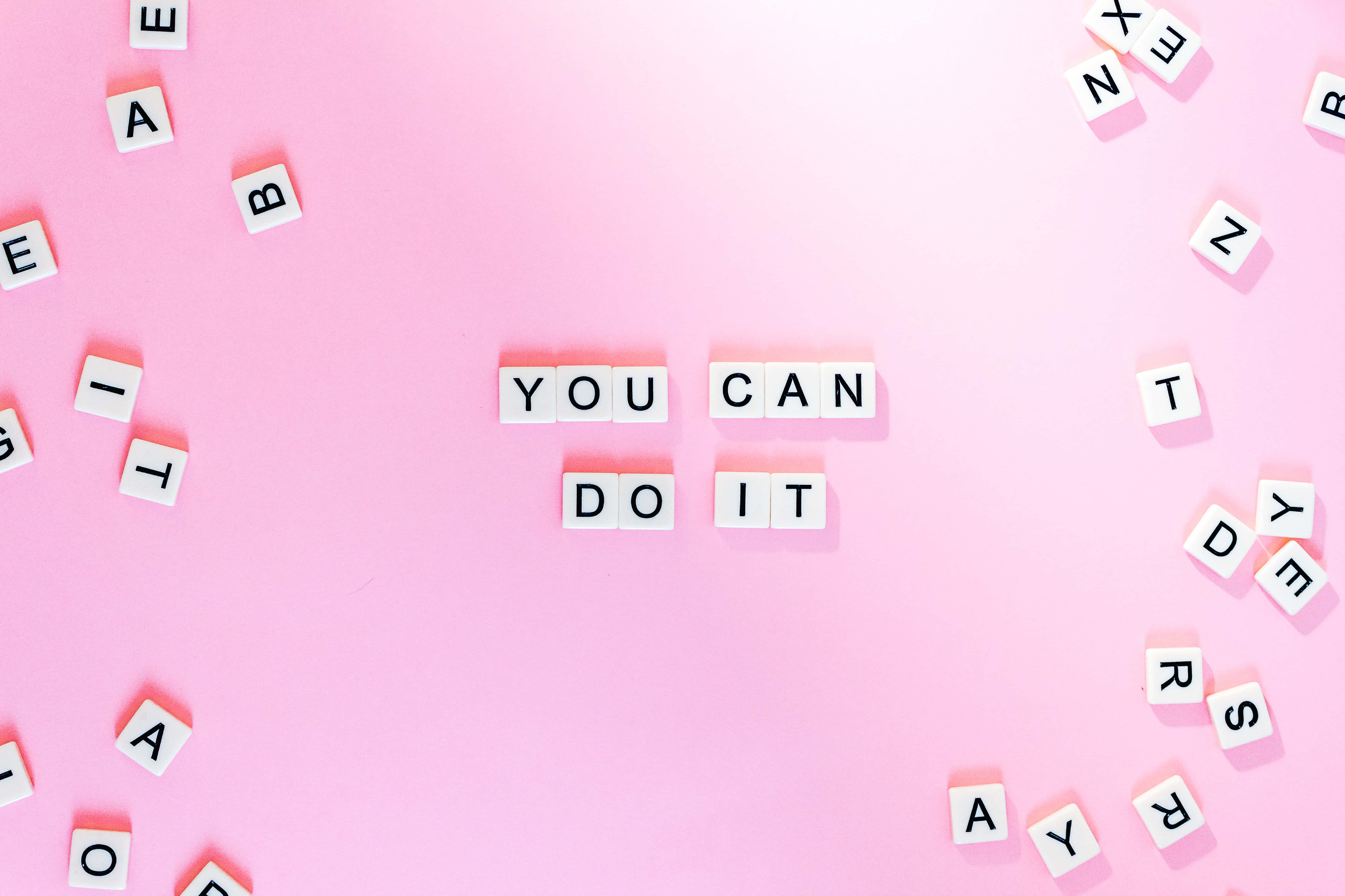 桌布 壁紙 — 白色字母瓷磚在柔和的粉紅色背景中央拼出「YOU CAN DO IT」，周圍散落著字母。