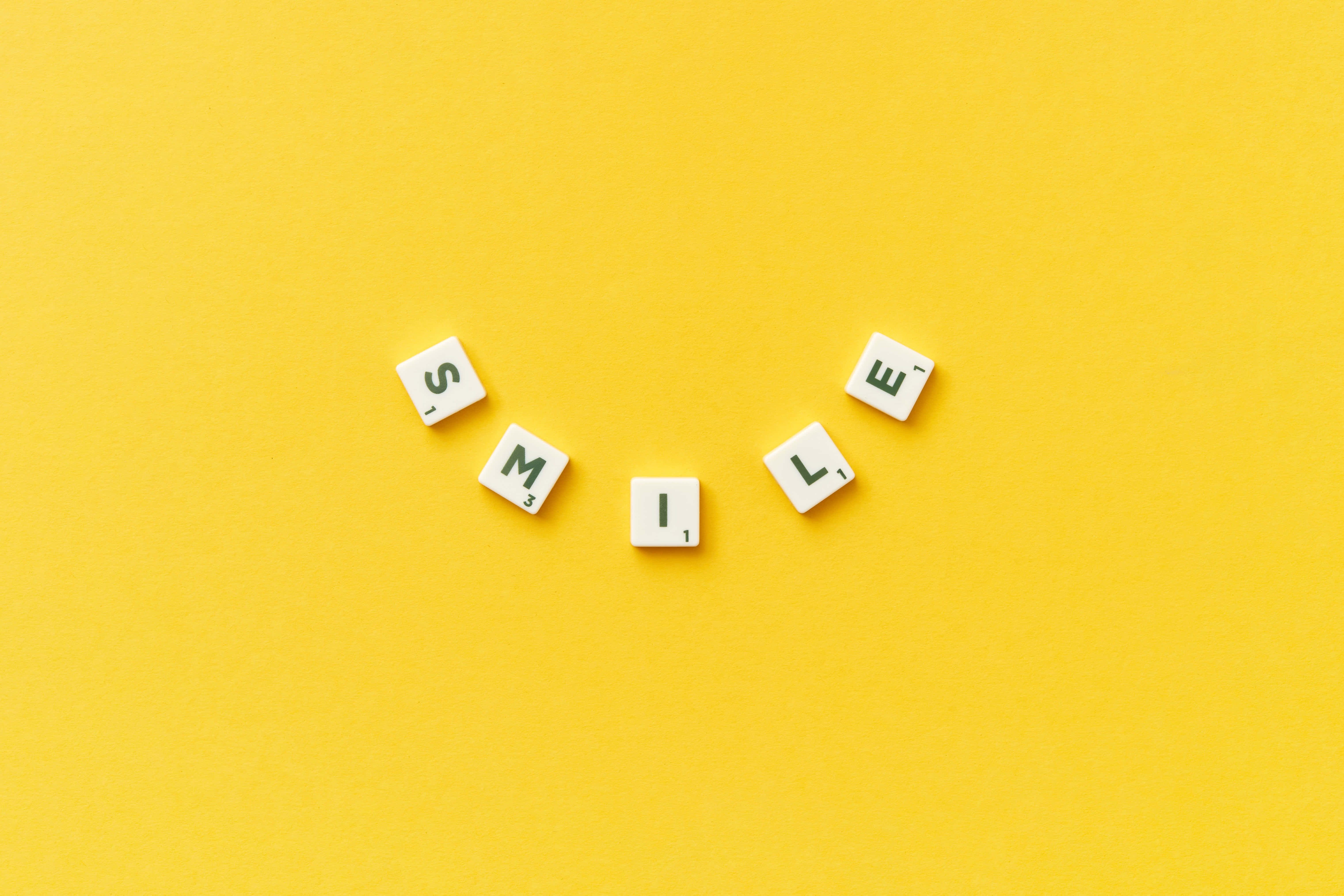 Smile 桌布 壁紙 — 白色字母瓷磚拼出 'SMILE'，以彎曲微笑形狀排列在鮮豔的黃色背景上。