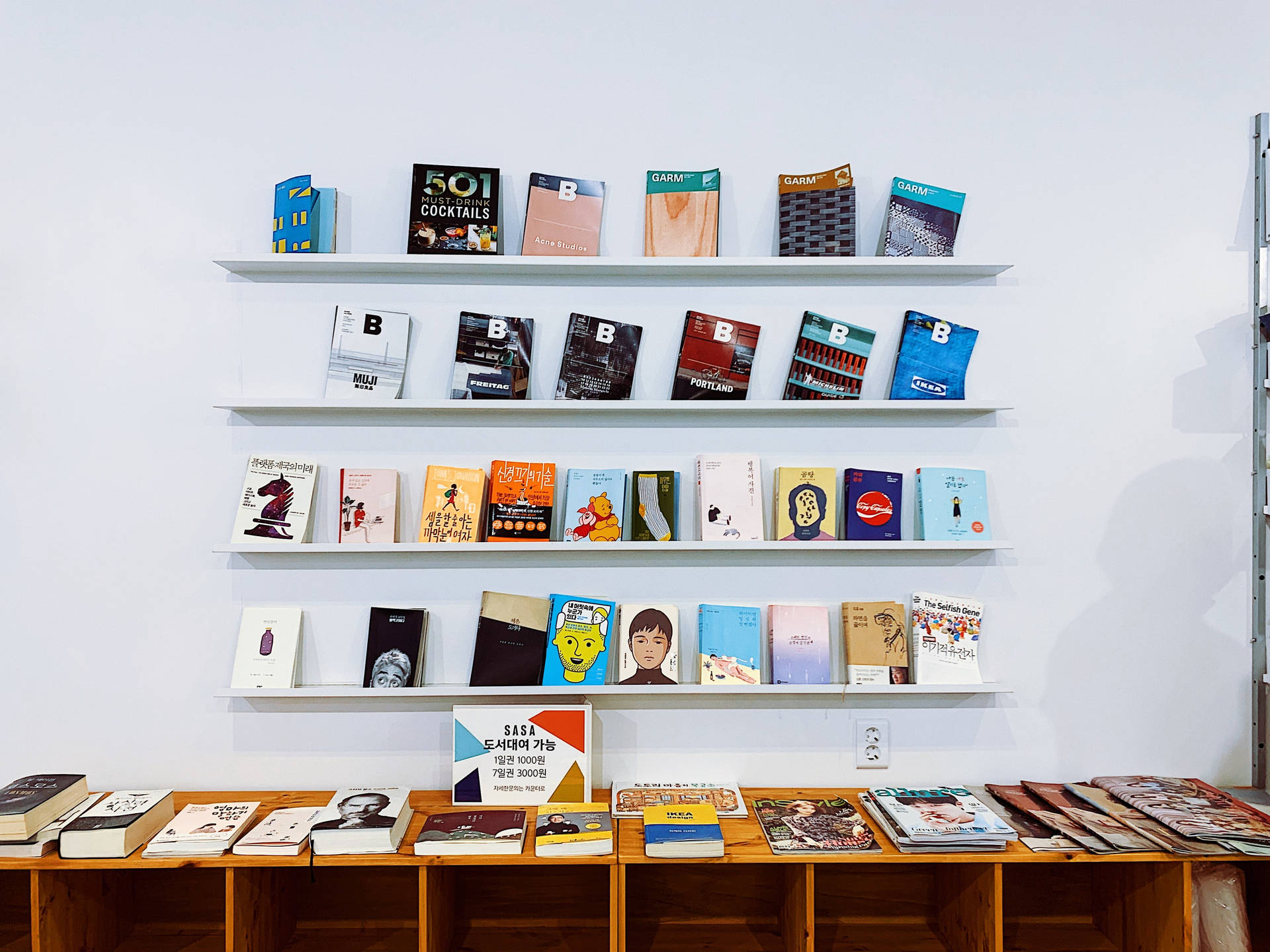 書本 桌布 壁紙 — 白色層架上展示色彩繽紛的書籍，背景為素色牆壁，下方木桌上亦放有更多書籍。
