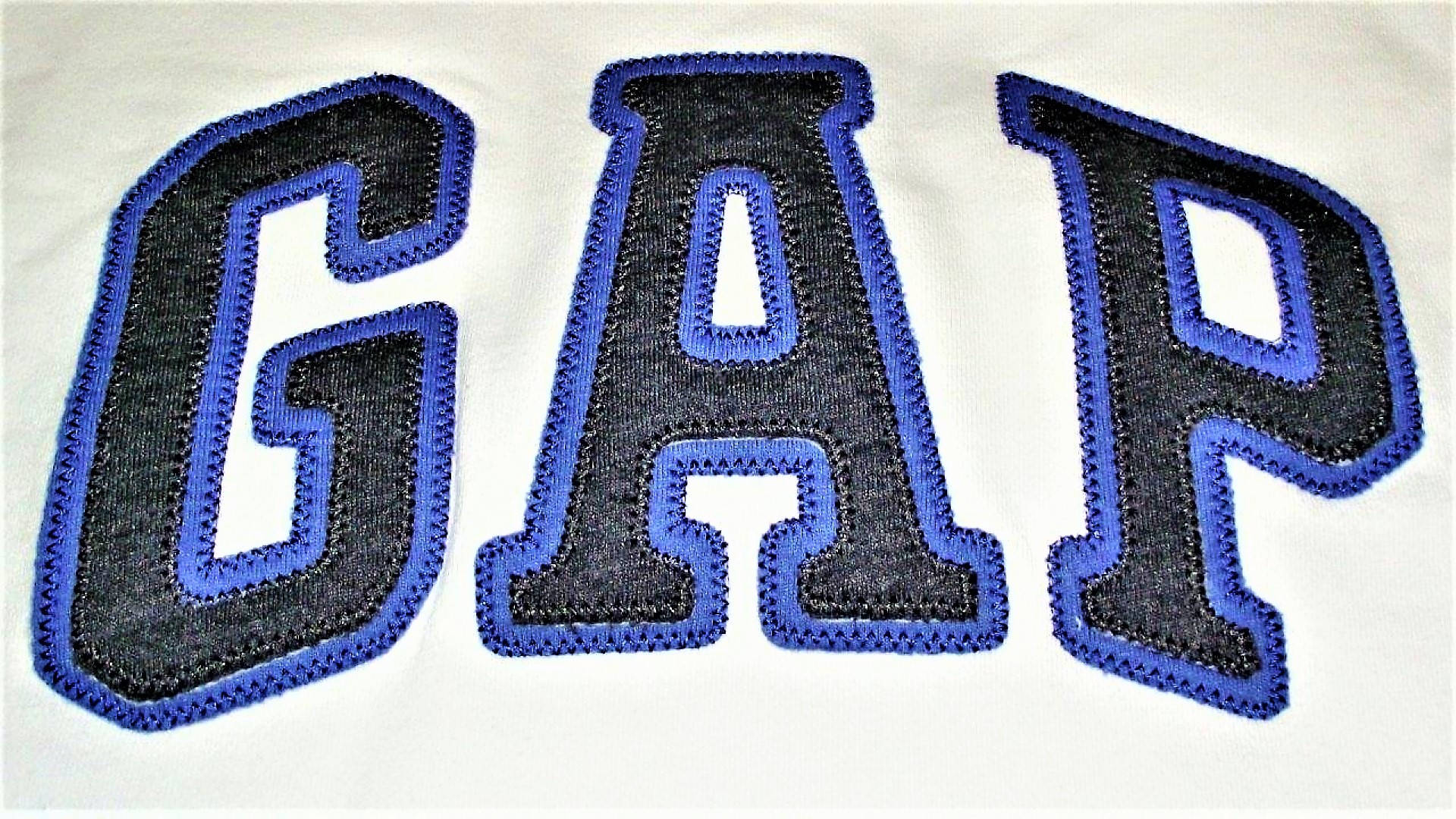 Gap 桌布 壁紙 — 白色布料上大膽黑色和藍色刺繡GAP標誌的特寫，可見縫線質感。