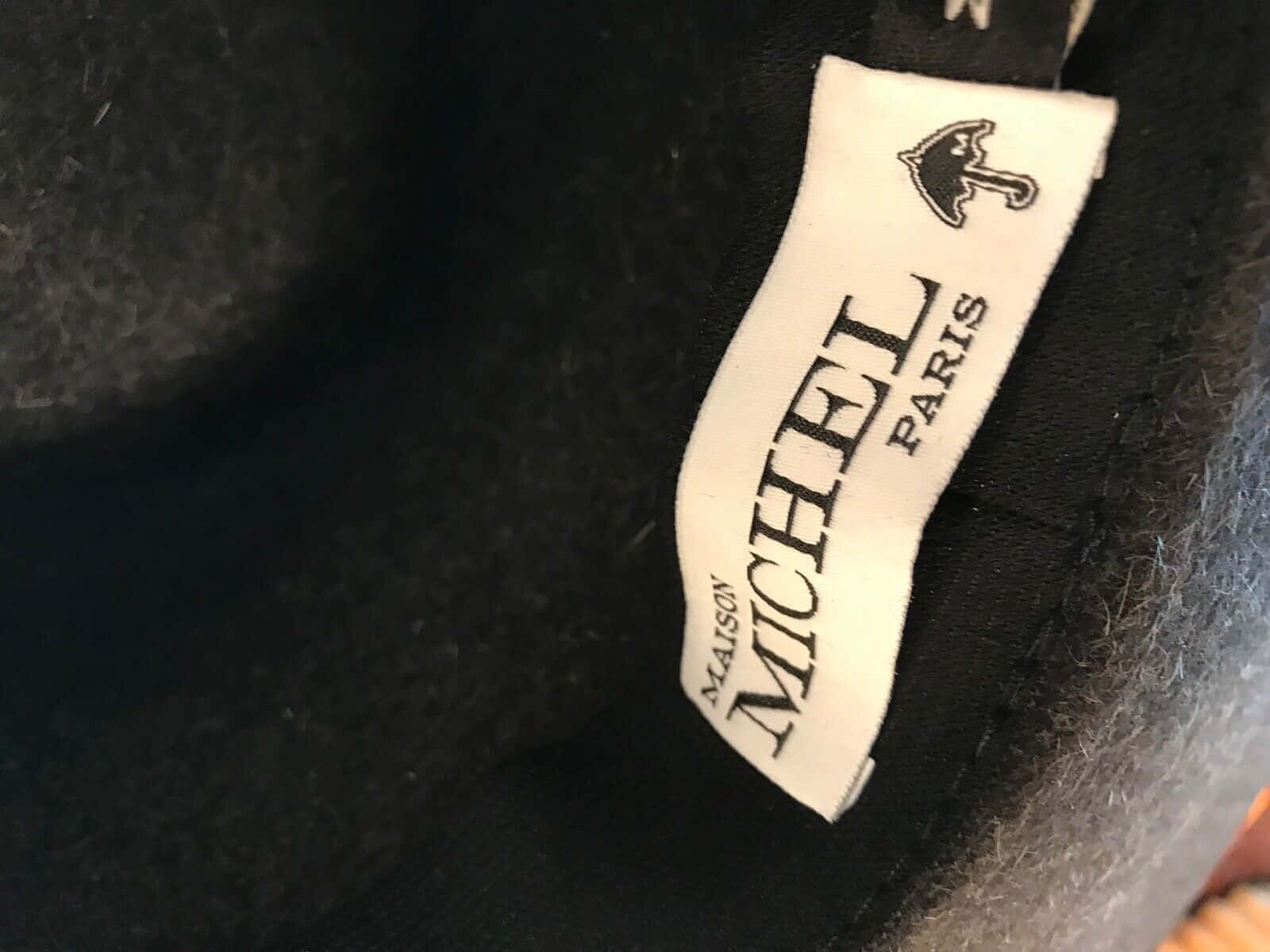Clothes 桌布 壁紙 — 白色布料標籤的特寫，上面寫著 Maison Michel Paris，並帶有縫在深色紋理羊毛上的雨傘標誌。
