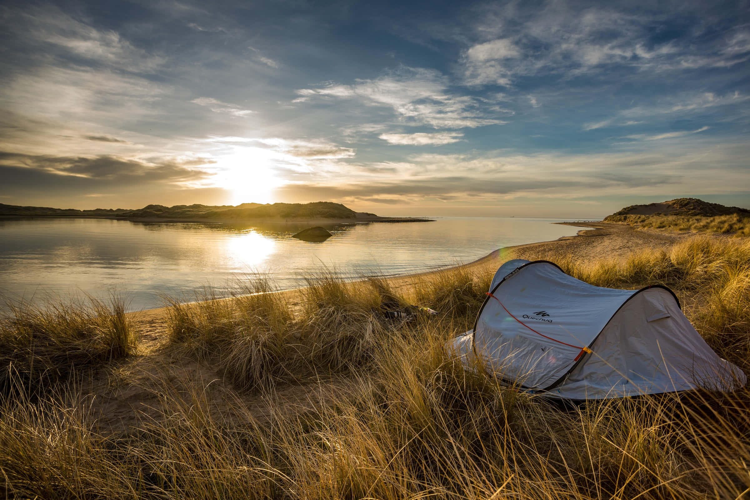 Camping 桌布 壁紙 — 白色帳篷坐落在金色海灘草叢中，夕陽西下，溫暖的光線反射在平靜的水面和遠處的山丘上。