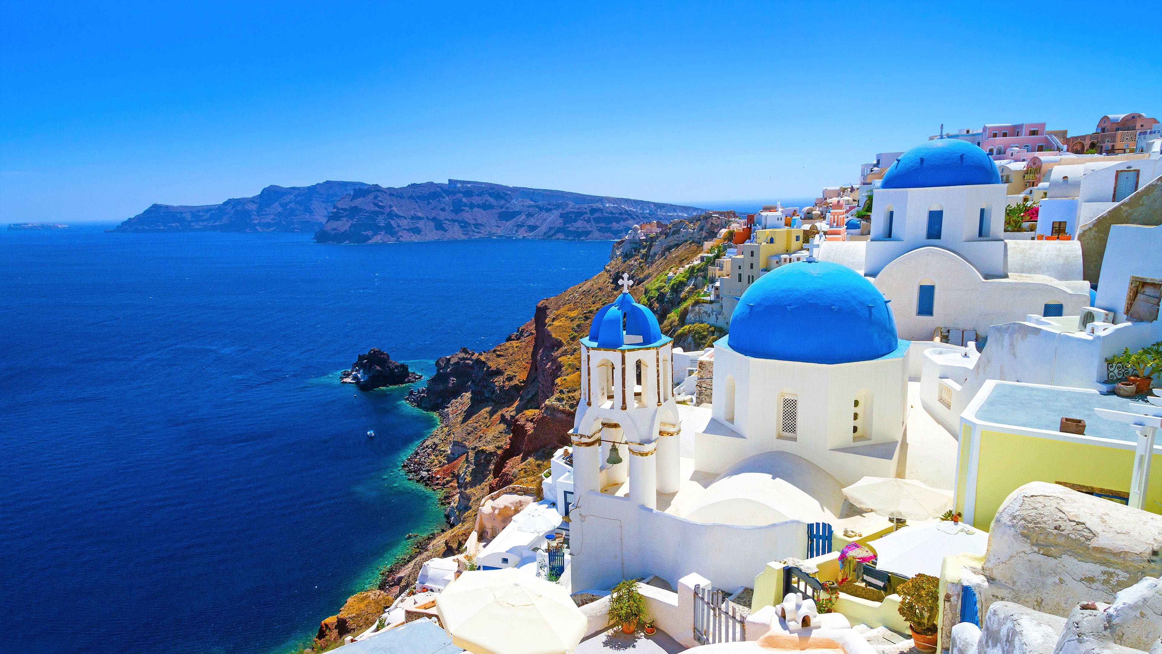 Santorini 桌布 壁紙 — 白色建築物搭配充滿活力的藍色圓頂，戲劇性地棲息在懸崖上，俯瞰深藍色的愛琴海。