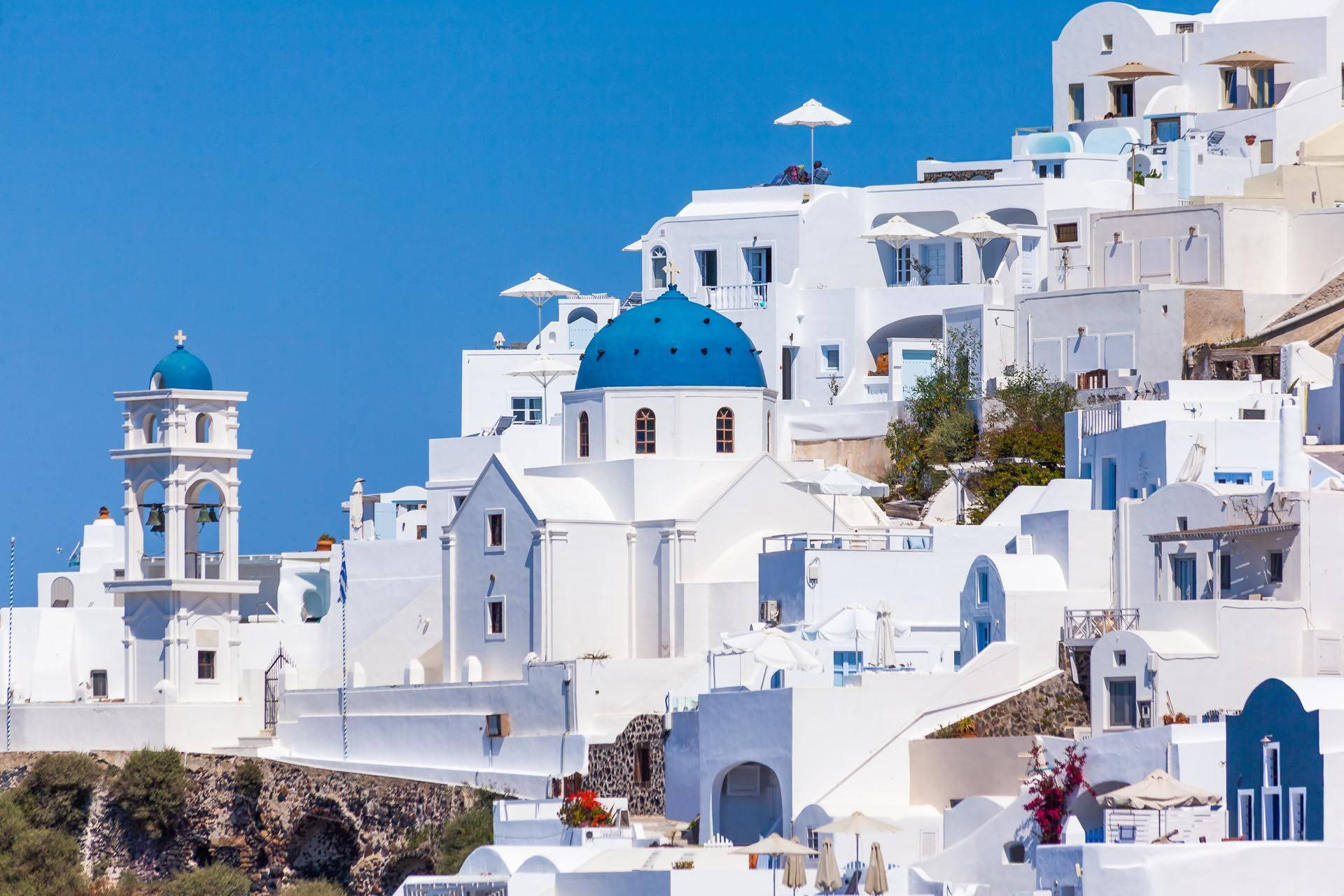 Santorini 桌布 壁紙 — 白色建築物搭配藍色圓頂和遮陽傘，在晴朗藍天下沿著陽光普照的懸崖層疊而下。