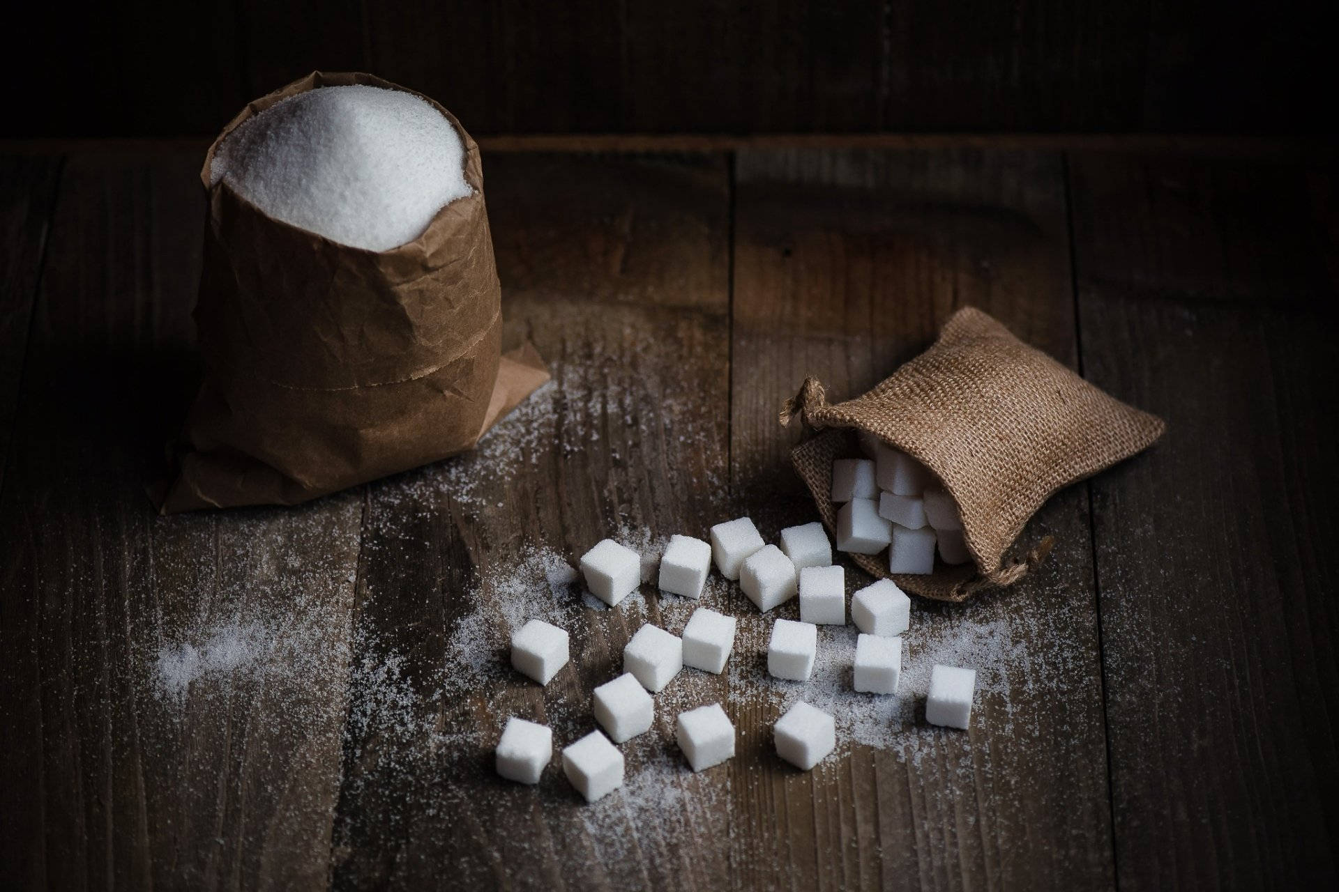 Sugar 桌布 壁紙 — 白色方糖和砂糖粒從麻布袋中灑落在深色木桌上，光線昏暗而富有戲劇性。