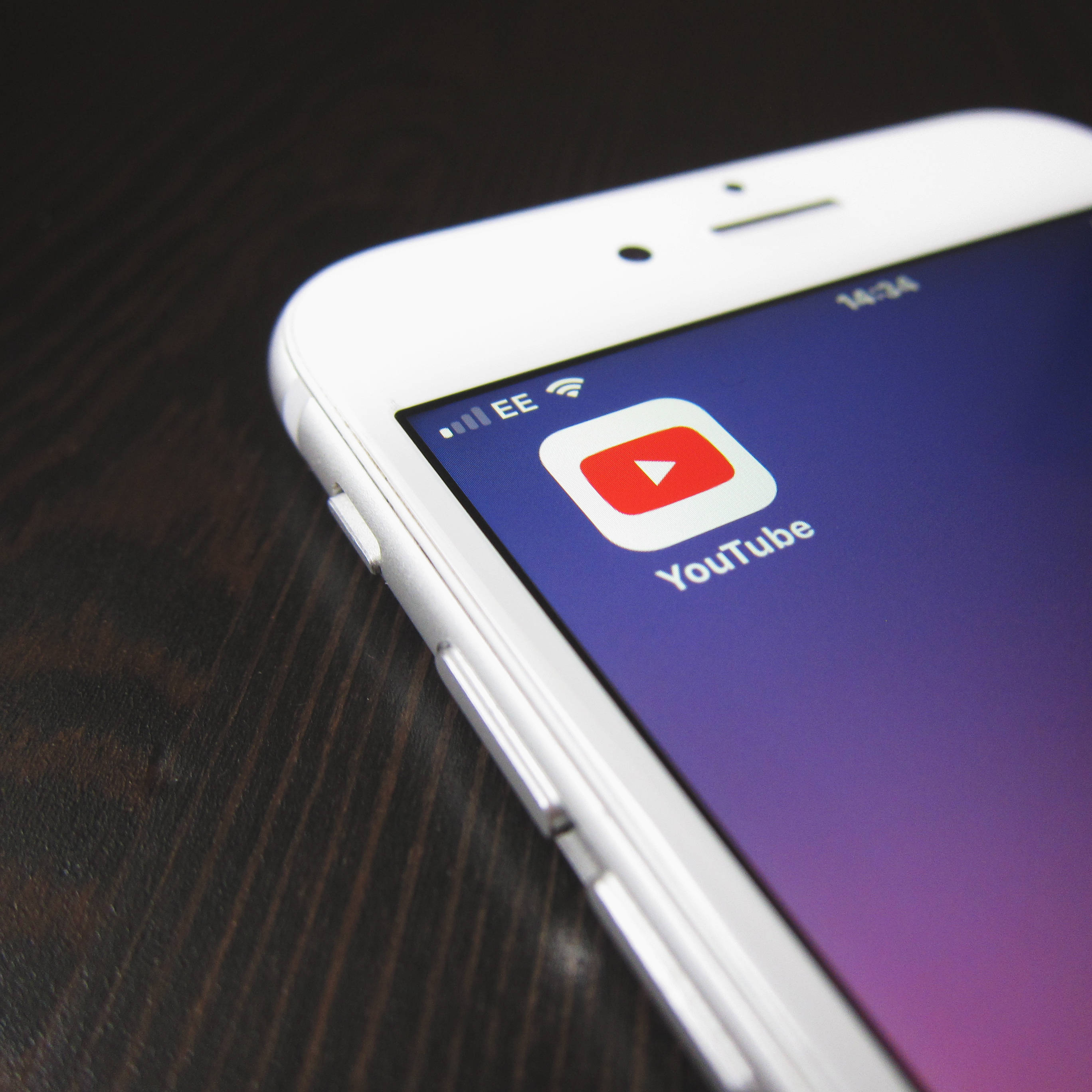 Banner 桌布 壁紙 — 白色智慧型手機的特寫，顯示紫色漸變螢幕上的 YouTube 應用程式圖示，放置在深色木質表面上。