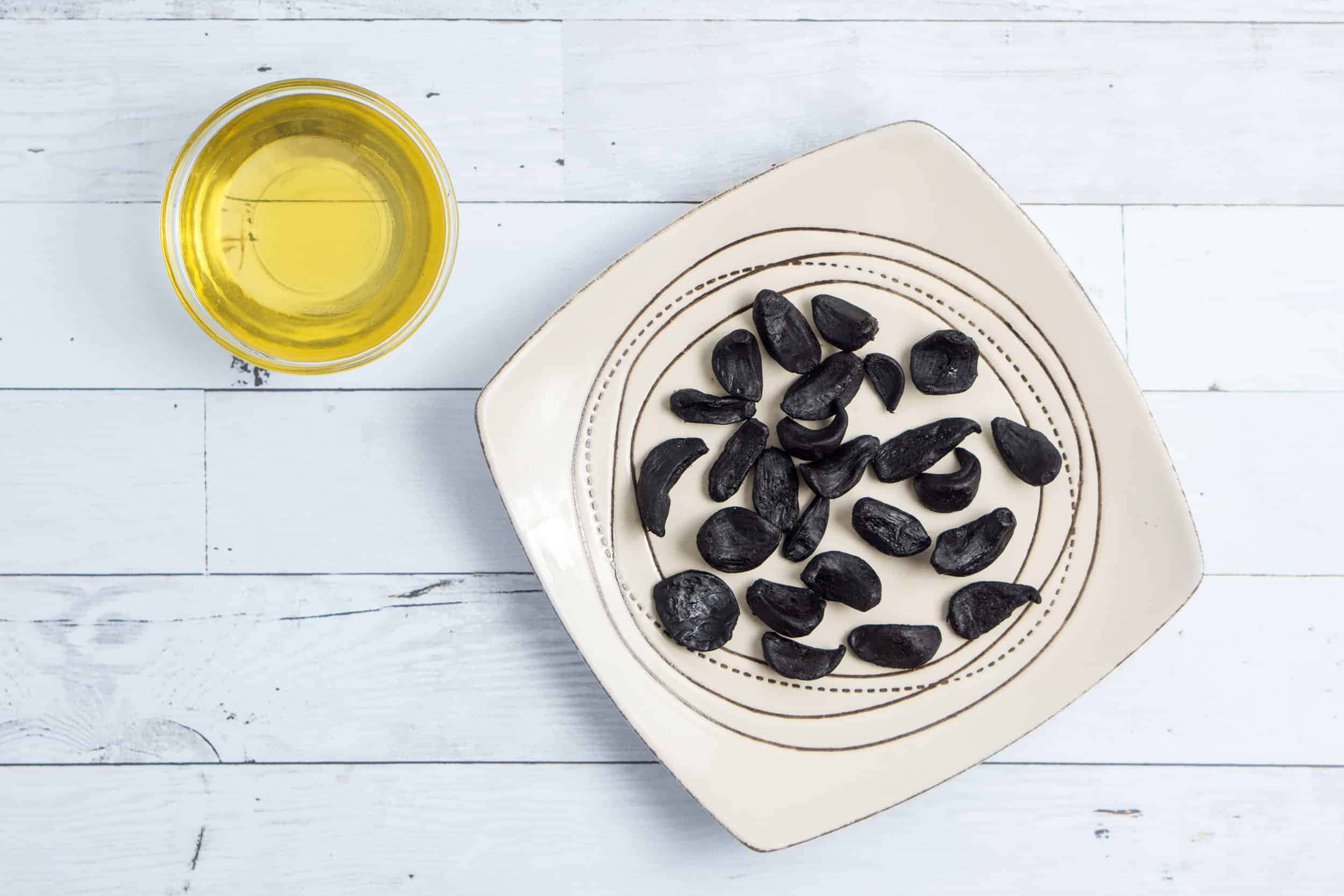 Black Garlic 桌布 壁紙 — 白色木質表面上，米色盤子上放著黑蒜瓣，旁邊有一杯金色油，從正上方俯瞰的視角。