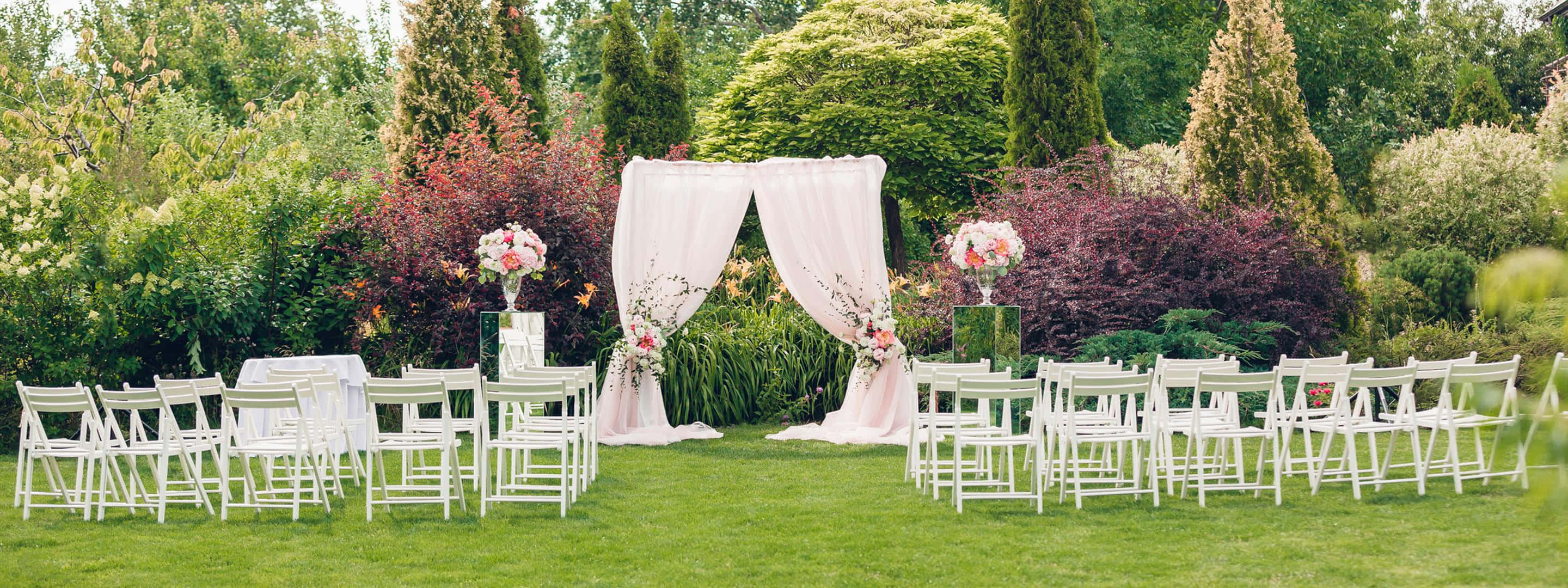 Garden Wedding 桌布 壁紙 — 白色椅子在綠色草地上排列成行，面向一個掛有布幔、飾有鮮花並兩旁有鮮豔灌木的拱門。